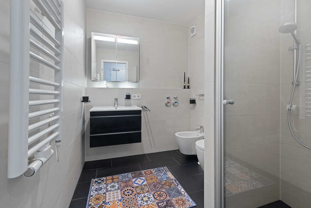 Modernes Badezimmer mit Dusche, Waschtisch, Bidet, WC und buntem Teppich auf schwarzem Boden.