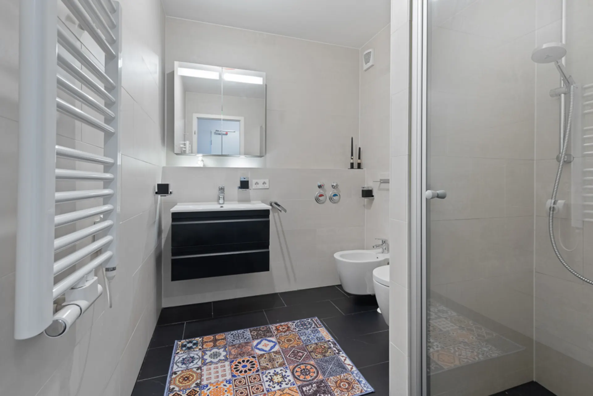 Modernes Badezimmer mit Dusche, Waschtisch, Bidet, WC und buntem Teppich auf schwarzem Boden.