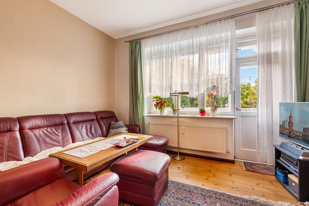 Wohnzimmer mit rotem Ledersofa, Holztisch, Pflanzen auf der Fensterbank und Fernseher.