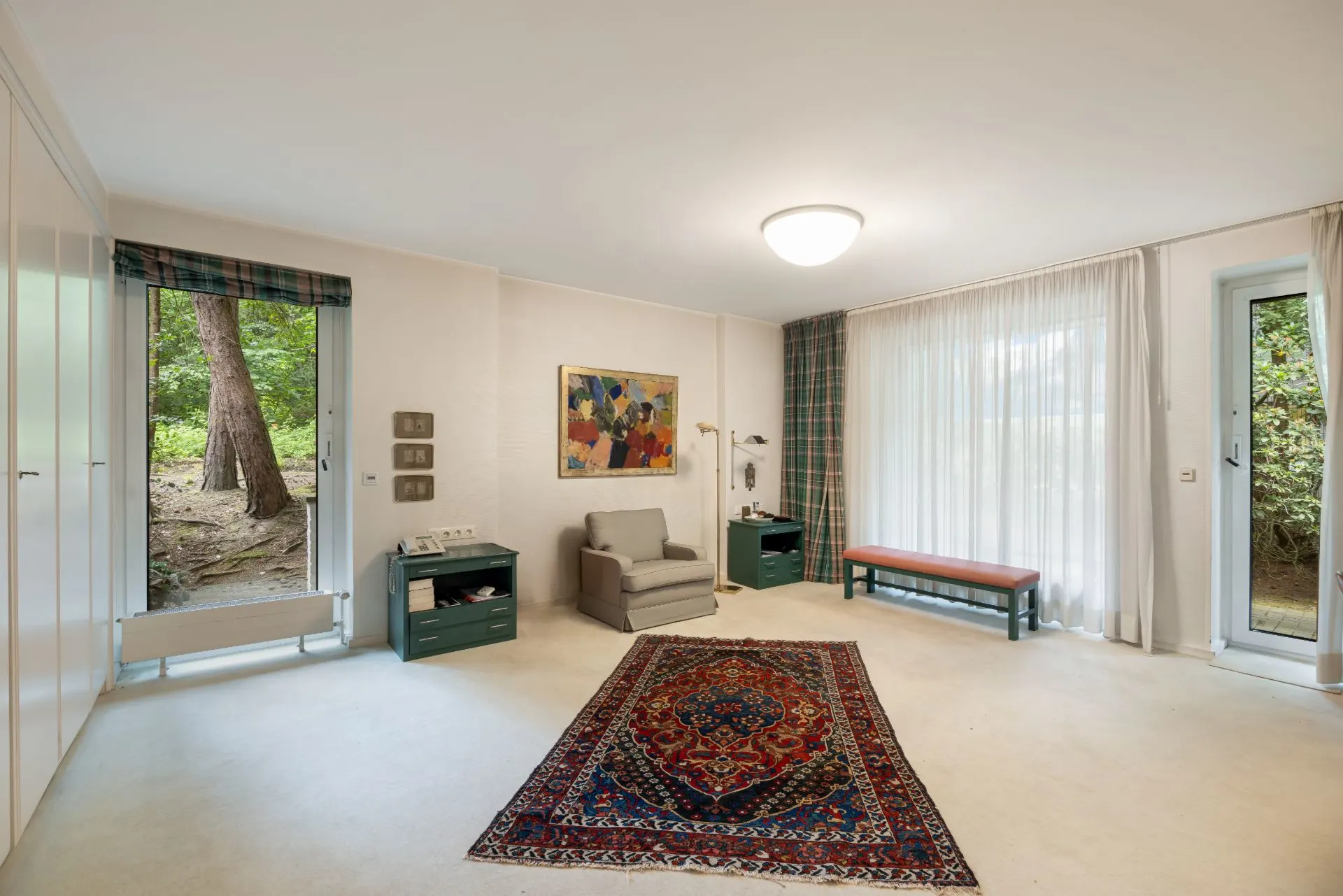 Helles Wohnzimmer mit Sessel, Teppich, Bank und großen Fenstern mit Blick ins Grüne.