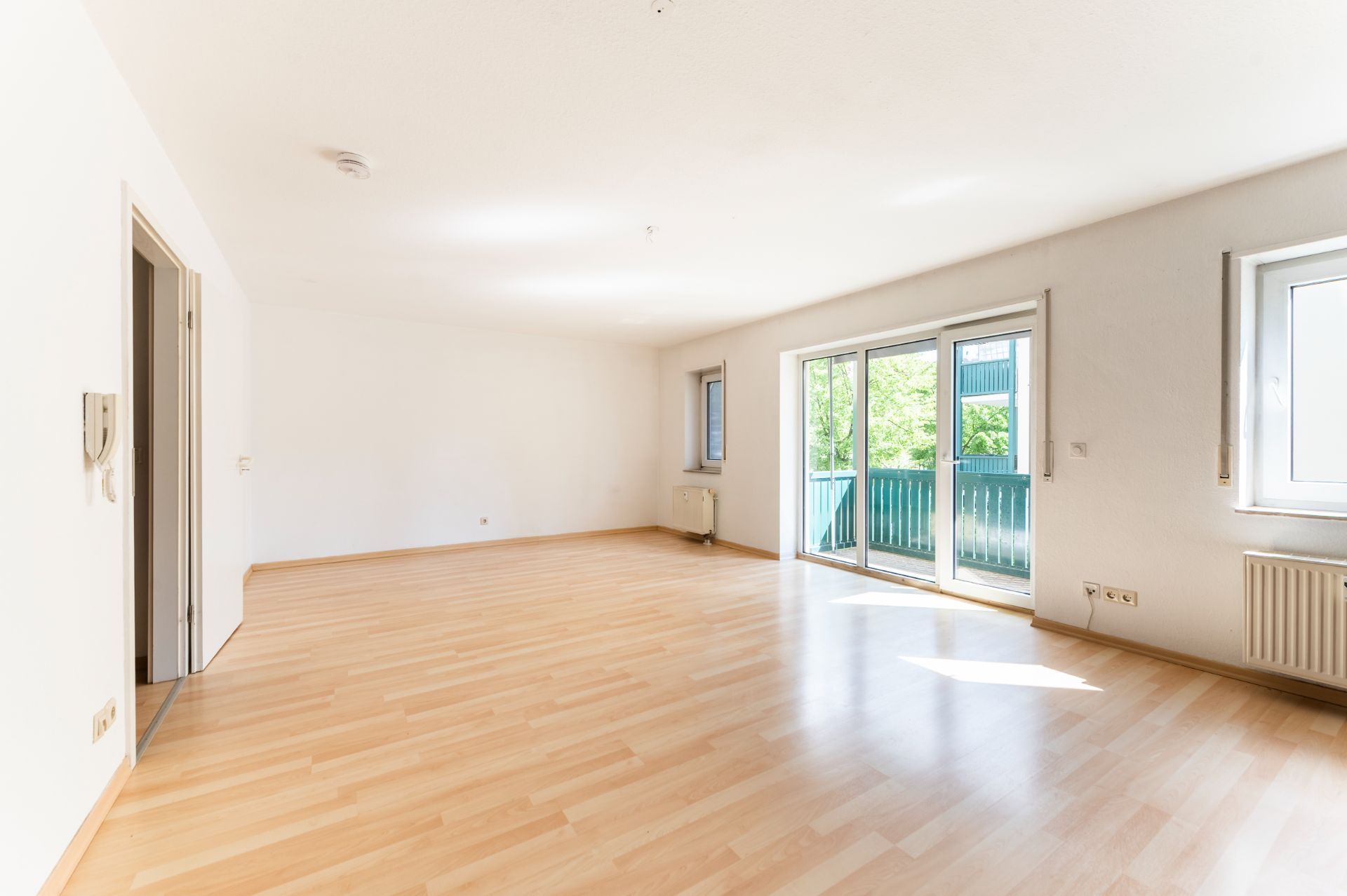 Heller, leerer Raum mit Parkettboden, weißen Wänden und großen Fenstern mit Zugang zu einem Balkon.