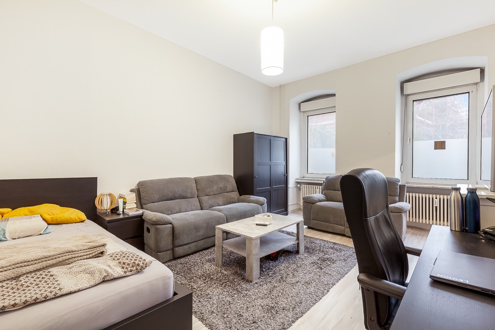 Helles Zimmer mit Bett, Sofa, Sessel, Schreibtisch und Couchtisch auf Teppich.