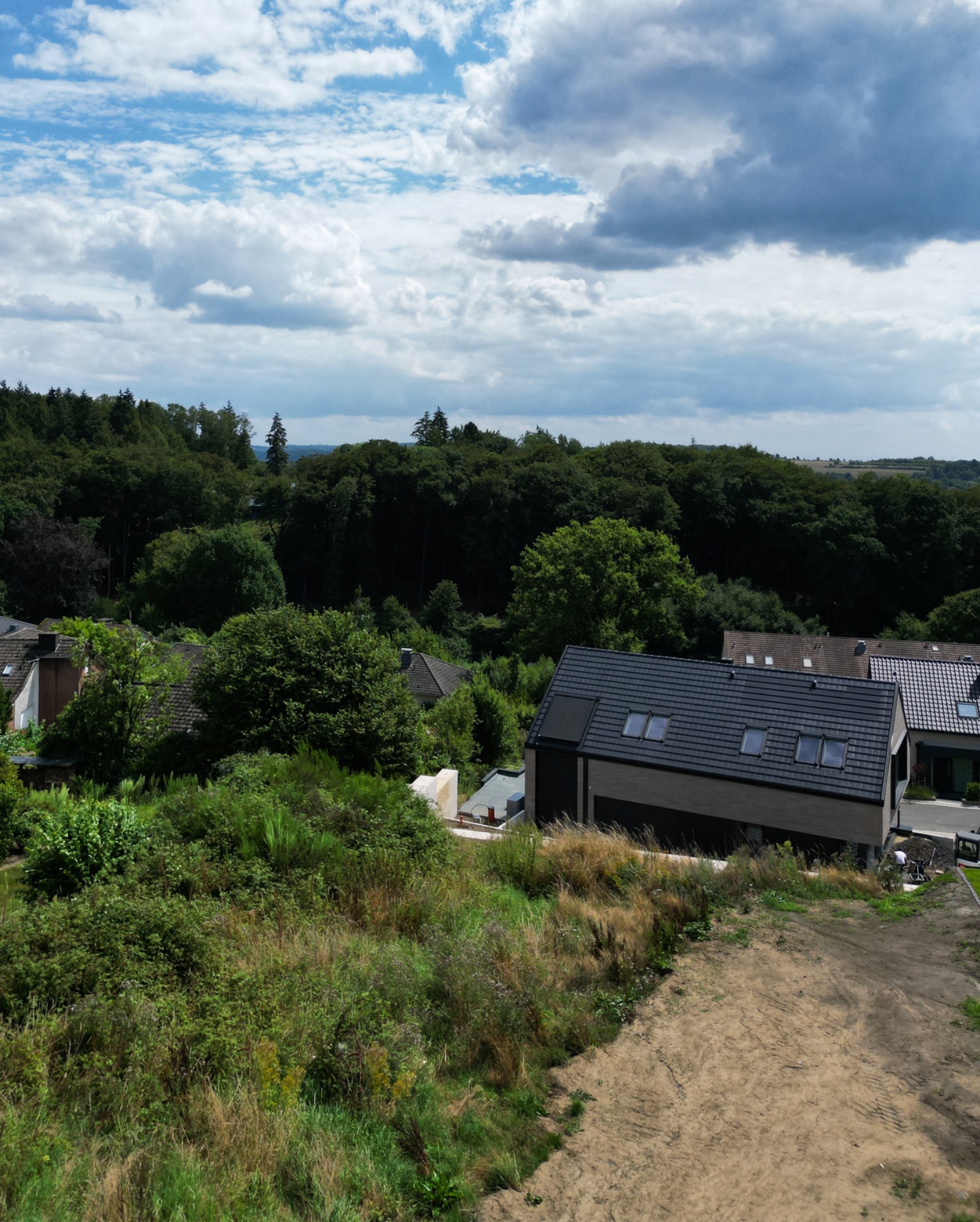 Haus mit dunklem Dach am Waldrand, davor ein Garten mit Wiese und kahlem Boden.