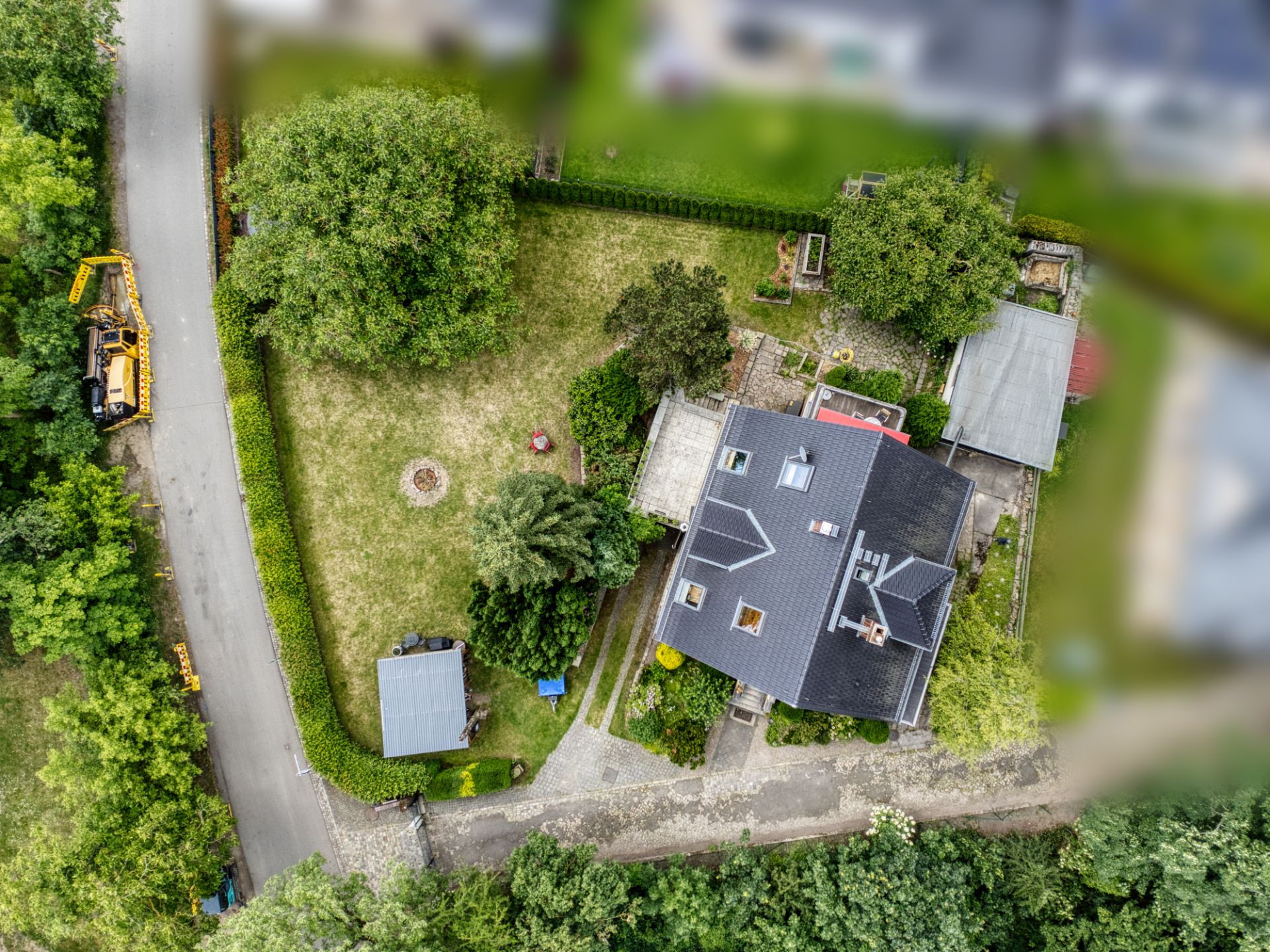 Luftaufnahme eines Hauses mit großem Garten, Bäumen und Nebengebäuden an einer Straße.