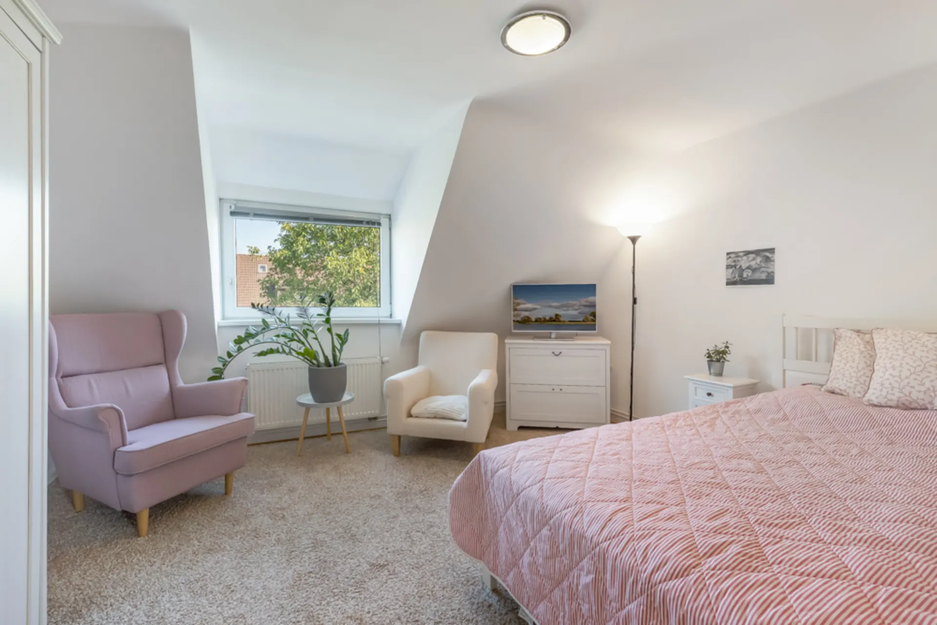 Helles Schlafzimmer mit Doppelbett, zwei Sesseln, Fernseher, Pflanze und großem Fenster.