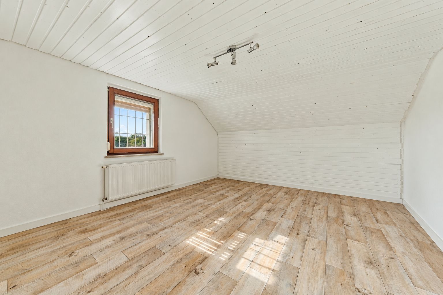 Helles, leeres Zimmer mit schräger Decke, Holzboden und einem Fenster mit Heizkörper.