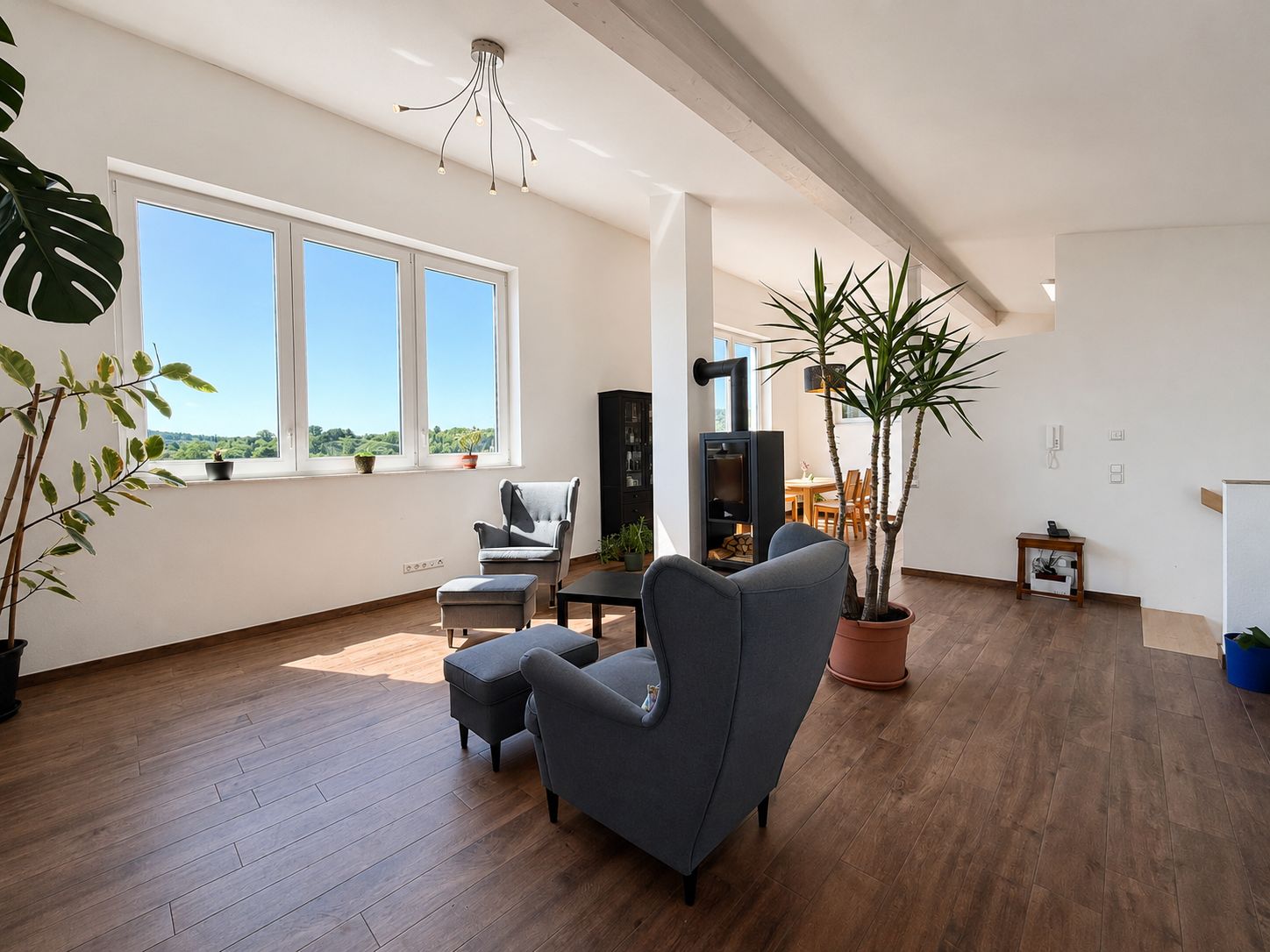 Helles Wohnzimmer mit großen Fenstern, Sessel, Couchtisch und mehreren Zimmerpflanzen auf Holzfußboden.