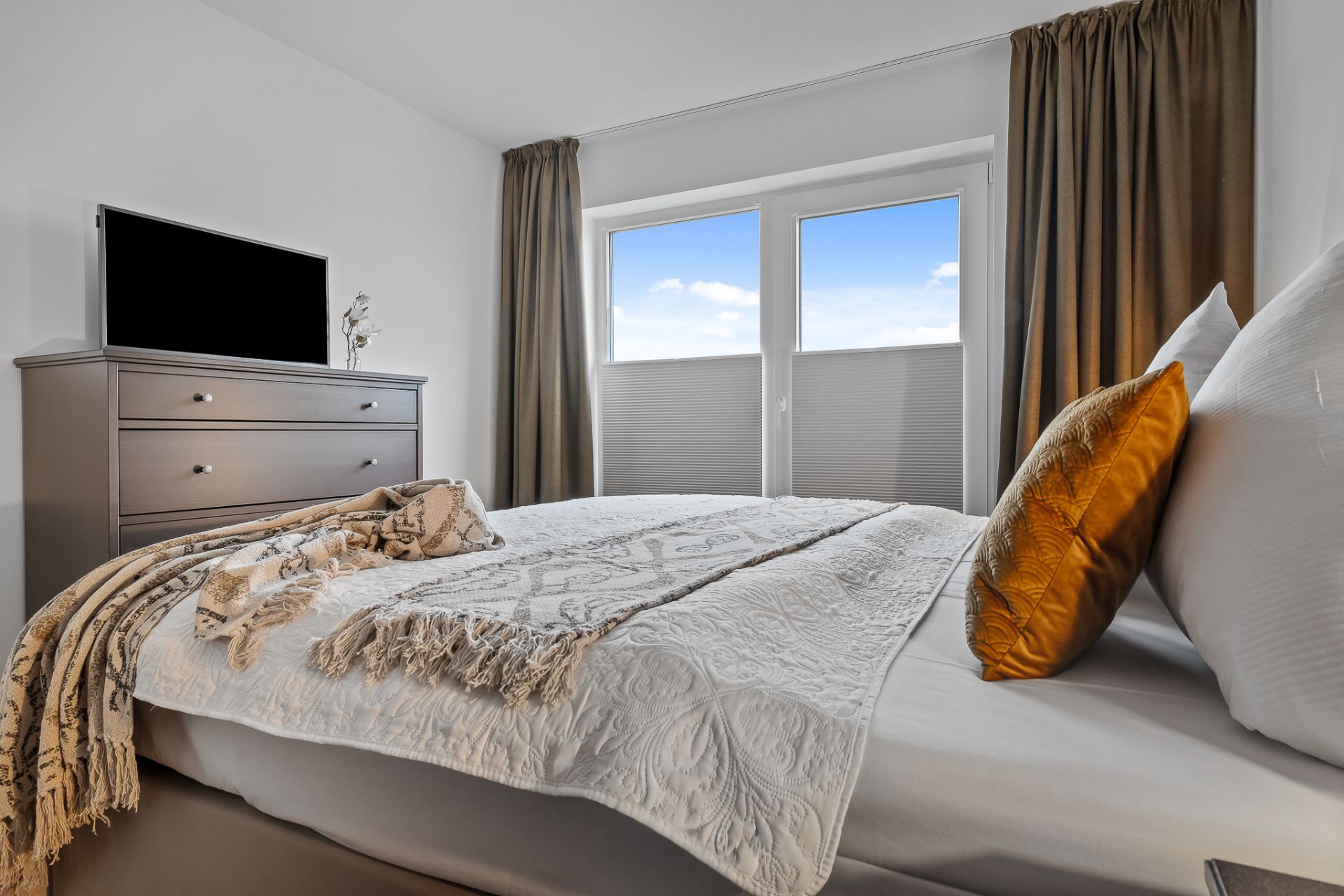 Schlafzimmer mit Bett, Kommode, Fernseher und großem Fenster mit Vorhängen.