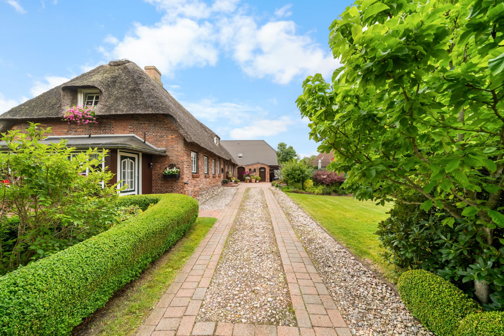 Backsteinhaus mit Reetdach, gepflasterte Einfahrt und gepflegter Garten mit Büschen und Bäumen.