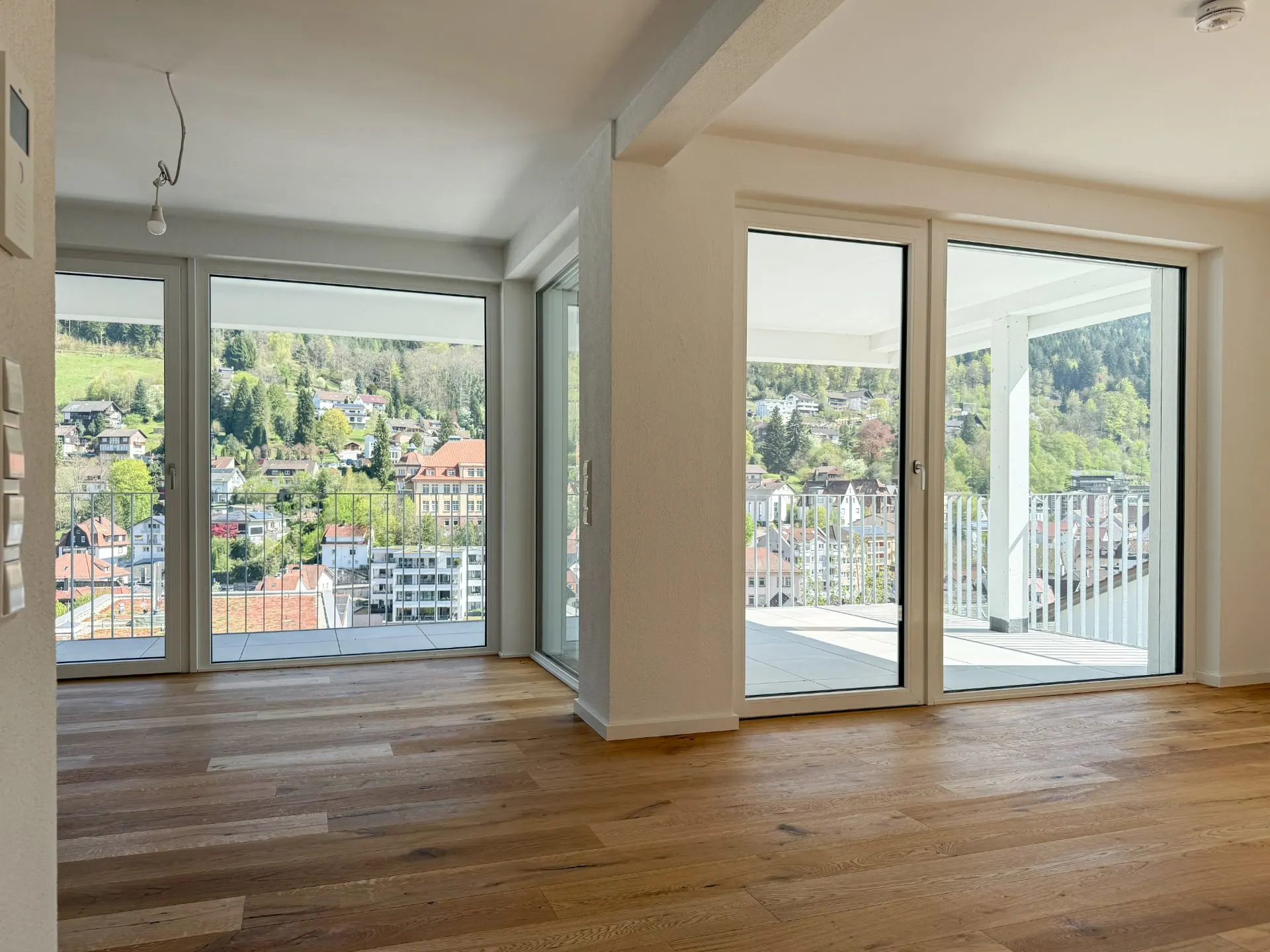 Heller Raum mit Parkettboden und großen Fenstern, Blick auf Balkon und grüne Hügellandschaft.