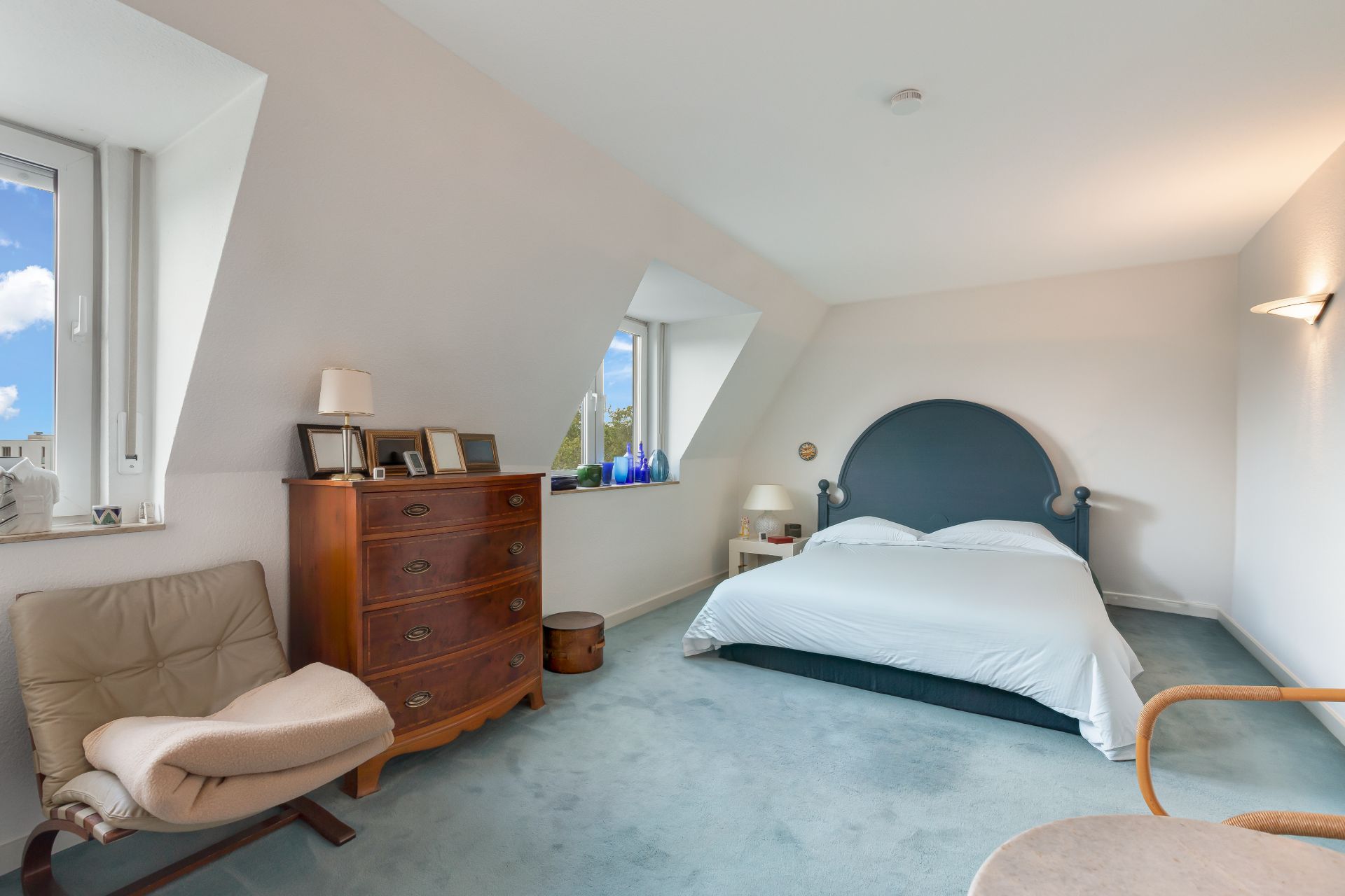 Helles Schlafzimmer mit Doppelbett, Kommode, Sessel und zwei Fenstern.