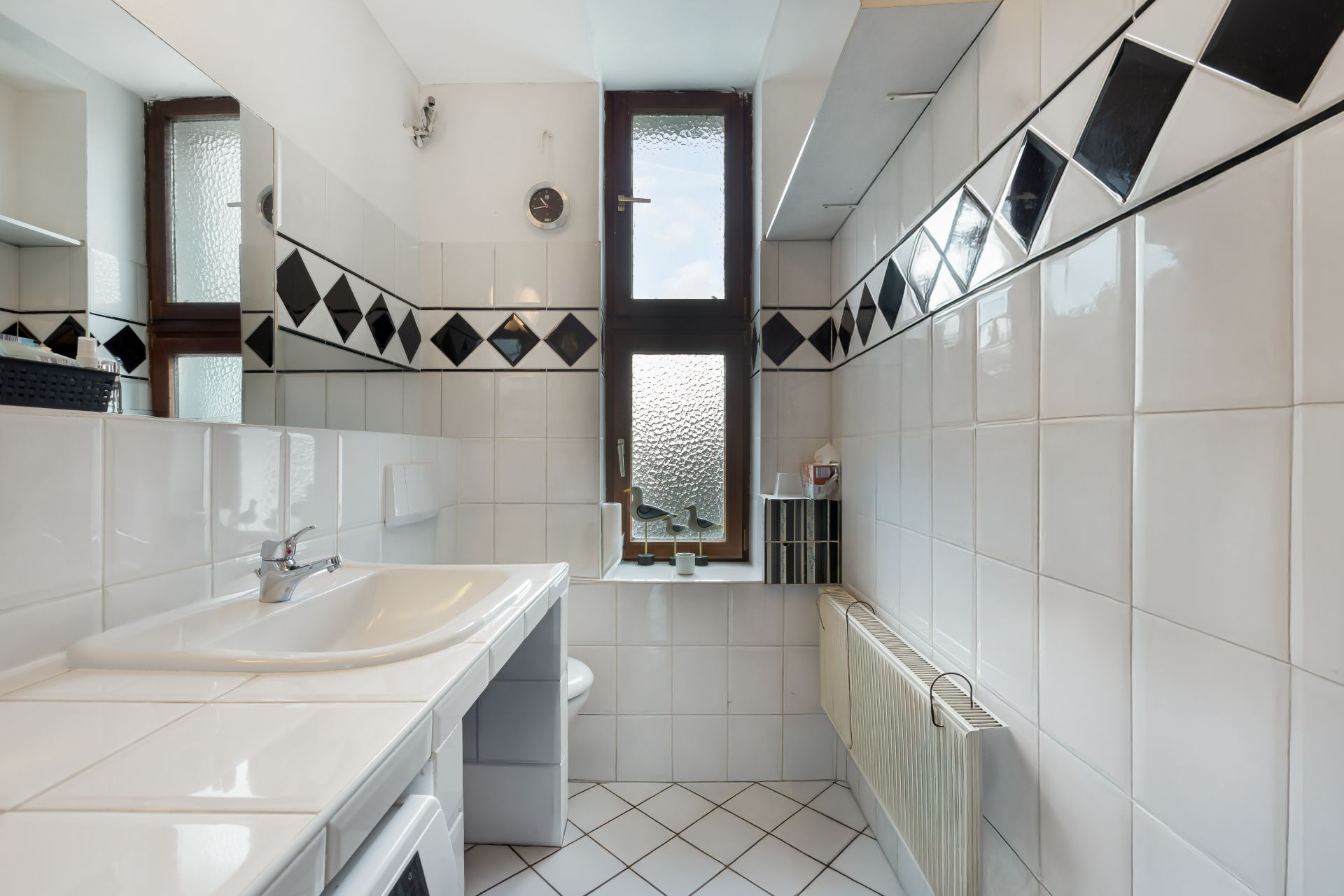 Helles Badezimmer mit weiß gefliesten Wänden, Waschbecken, Fenster und schwarzen Akzenten.