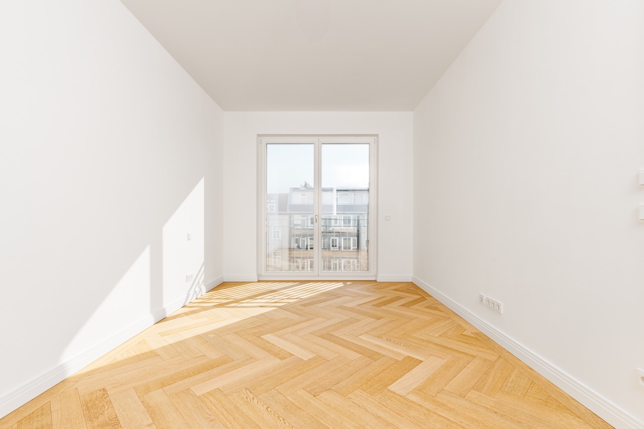 Helles, leeres Zimmer mit Fischgrätparkett und großem Fenster mit Blick auf andere Gebäude.