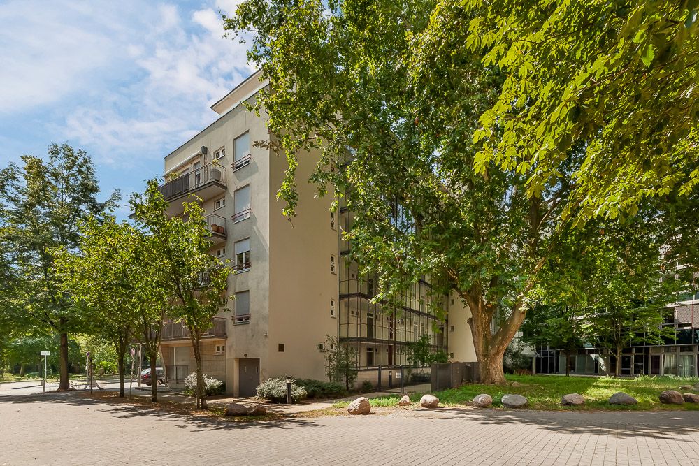 Mehrfamilienhaus mit Balkonen, umgeben von Bäumen und einem gepflasterten Weg.
