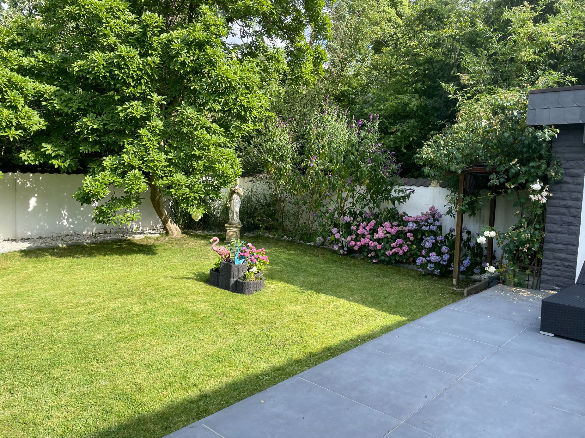 Garten mit Rasen, Baum, Blumenbeeten, einer Statue und einer Terrasse mit grauen Fliesen.