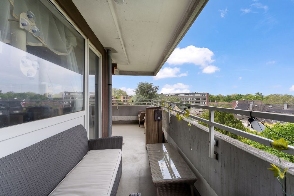 Balkon mit Sitzbank, Tisch und Blick auf Wohnhäuser und Bäume bei blauem Himmel.