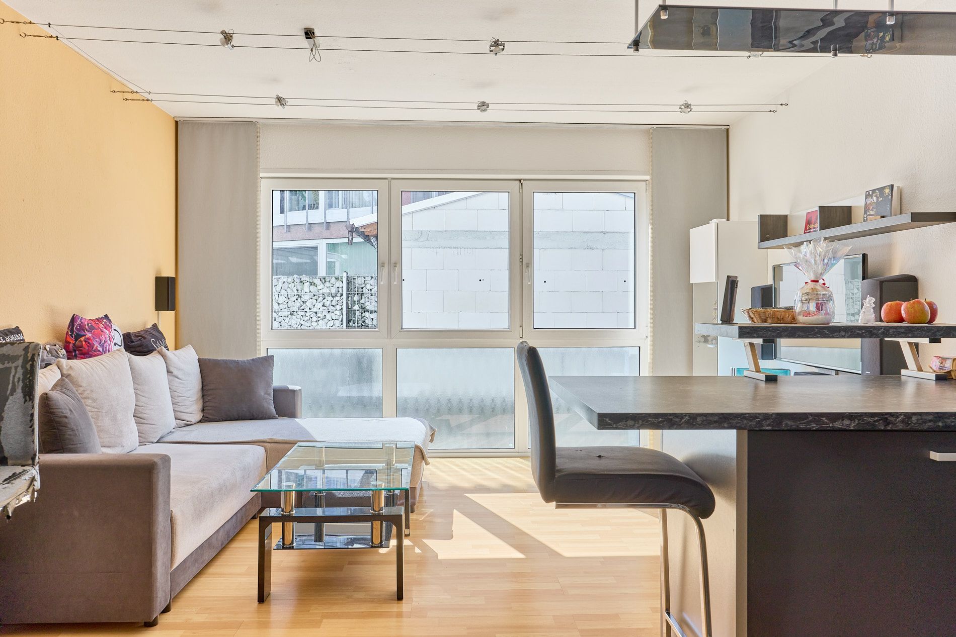 Helles Wohnzimmer mit grauem Sofa, Couchtisch, Barhockern und großen Fenstern.