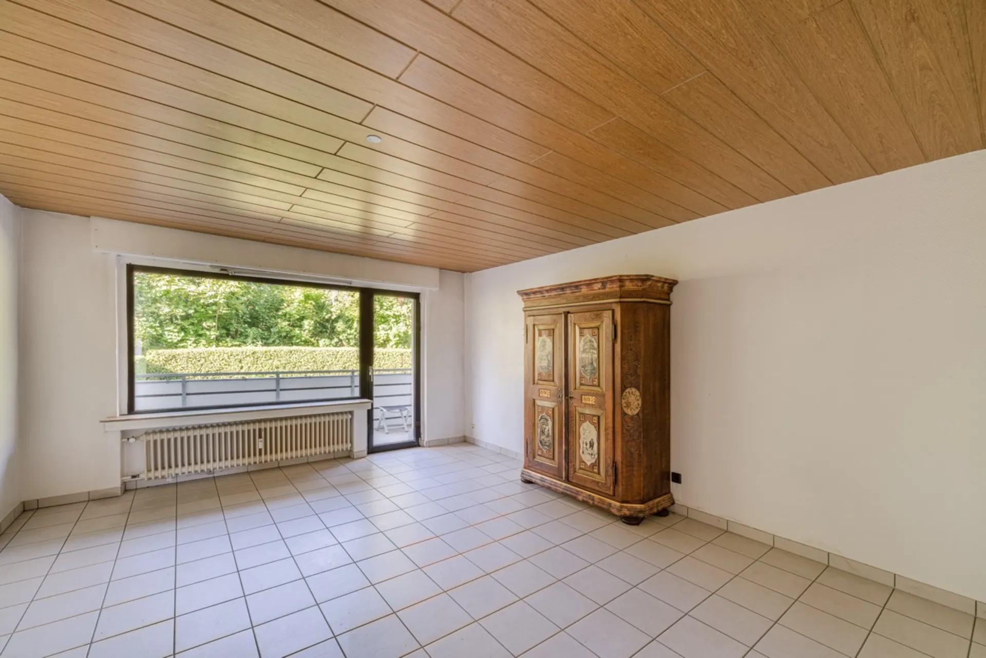 Helles Zimmer mit Fliesenboden, Holzdecke, großem Fenster und einem bemalten Holzschrank.