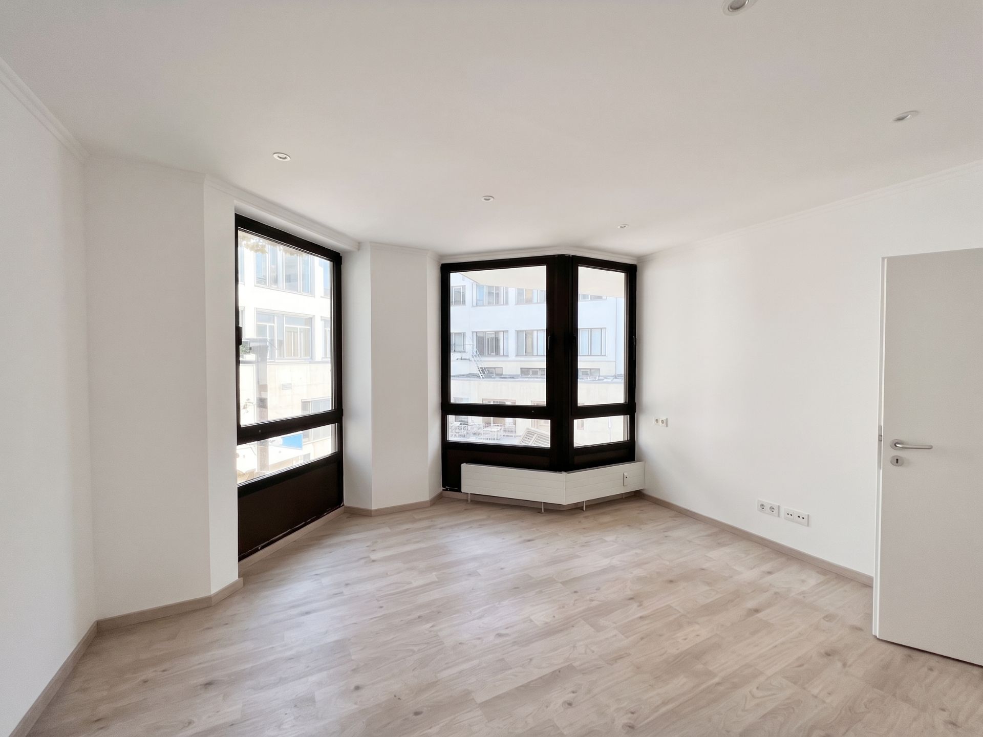 Heller leerer Raum mit großem Eckfenster, Holzfußboden und weißer Wand.