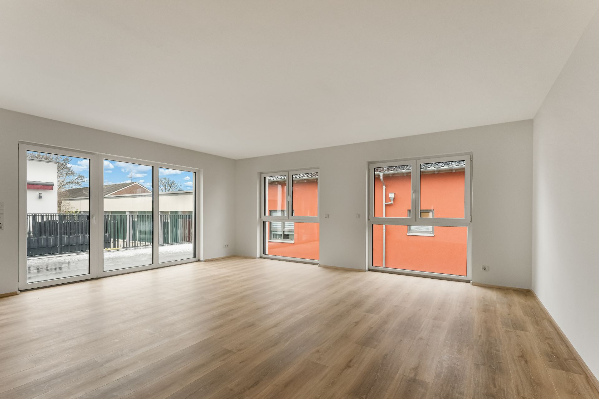 Helles, leeres Zimmer mit großen Fenstern und Parkettboden.