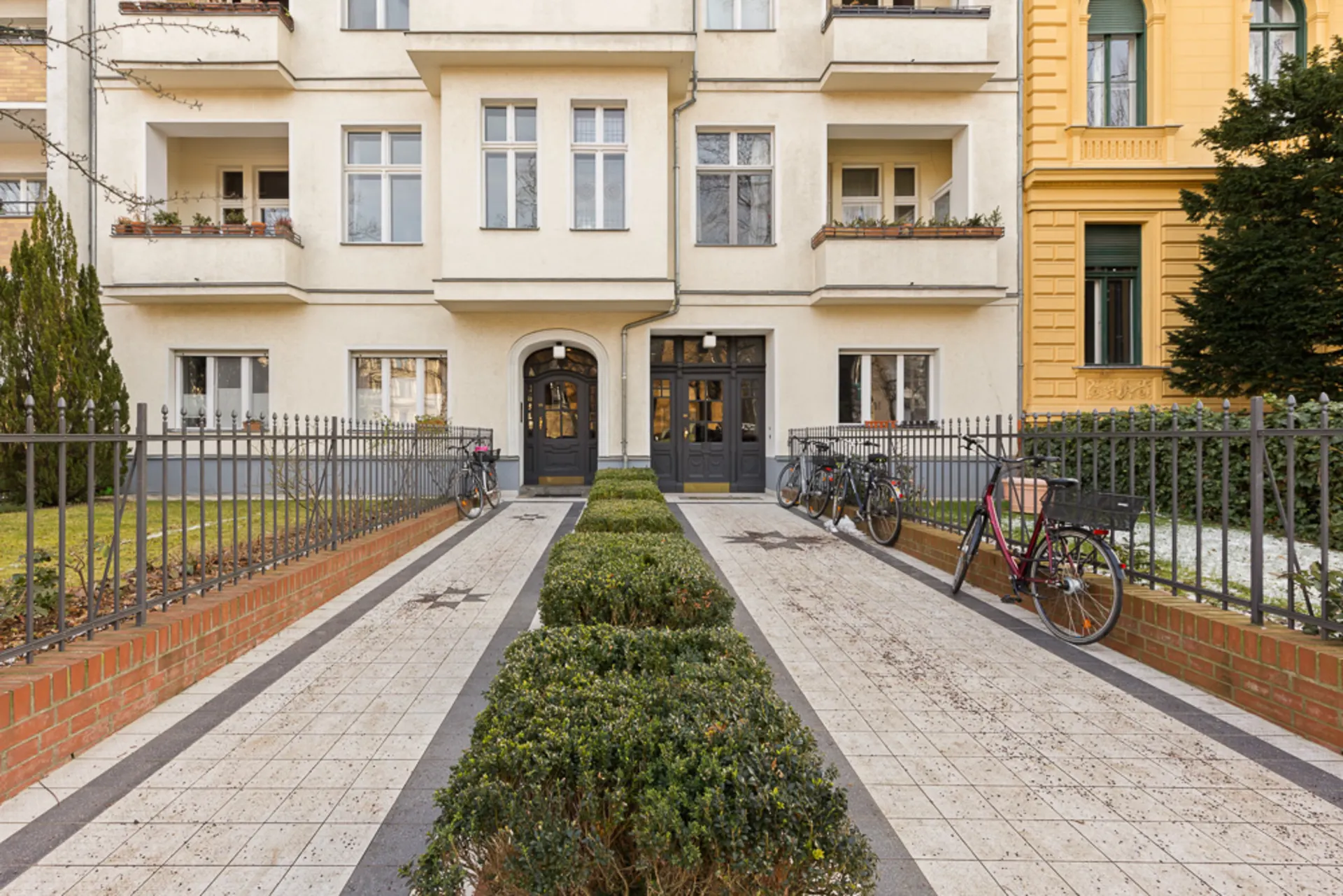 Eingang eines Wohnhauses mit Zweiwegen, Garten, Zaun und abgestellten Fahrrädern.