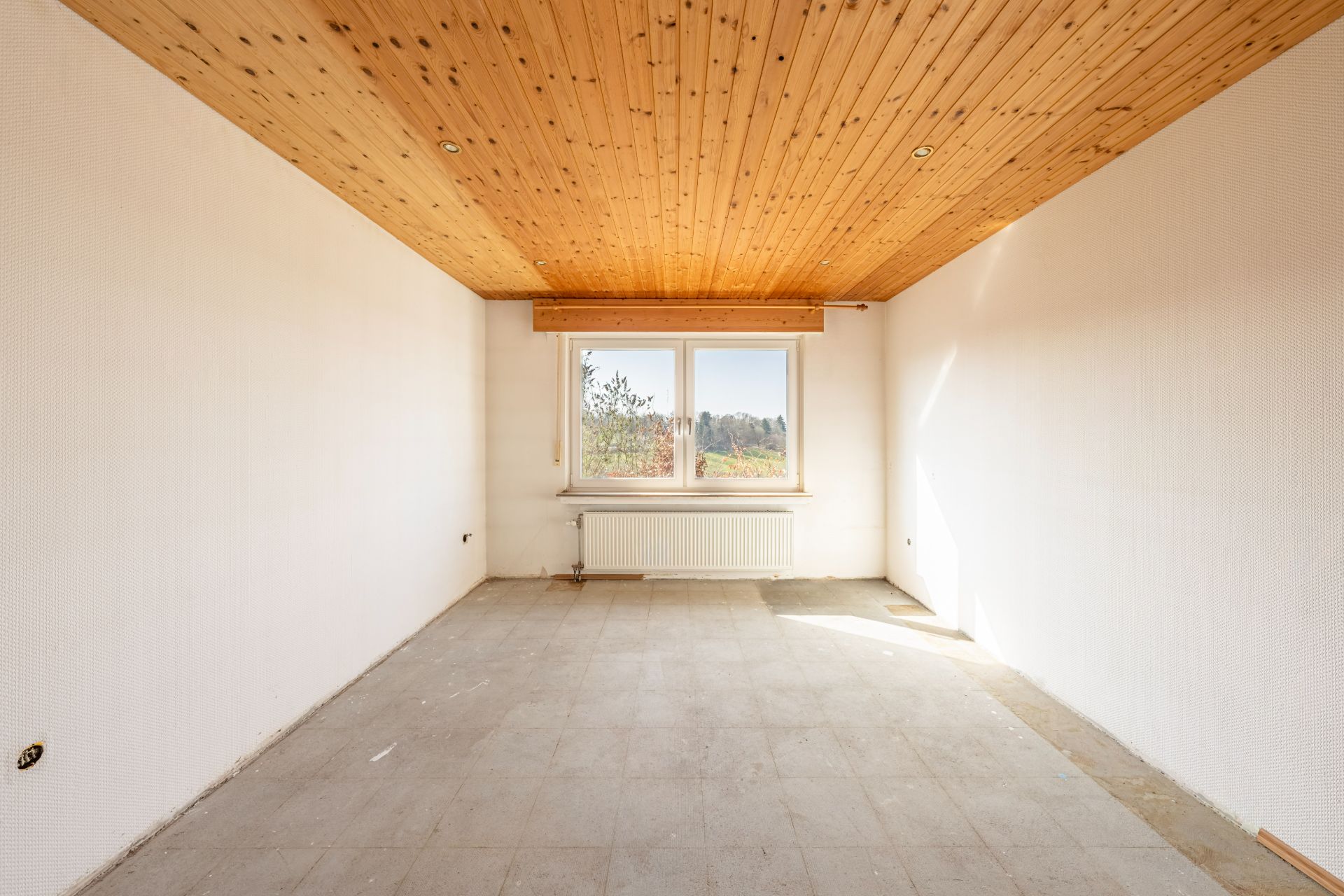 Leerer Raum mit Holzdecke, weißen Wänden, Fenster und Heizkörper, Fliesenboden.