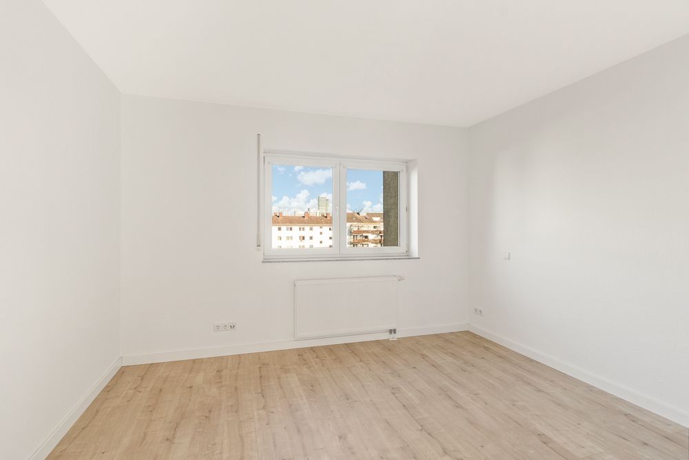 Leerer, heller Raum mit Holzfußboden, weißer Wand und Fenster mit Blick auf Nachbargebäude.