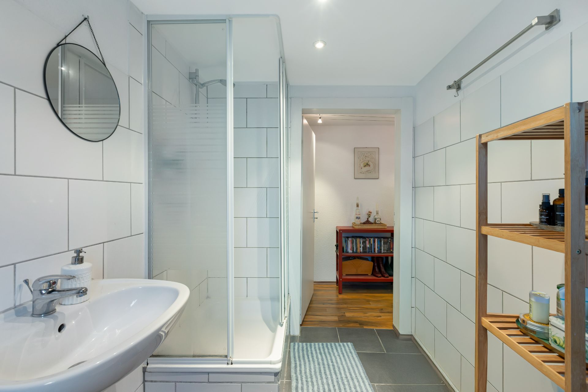 Helles Badezimmer mit Dusche, Waschbecken, Regal und rundem Spiegel, Fliesen an den Wänden.