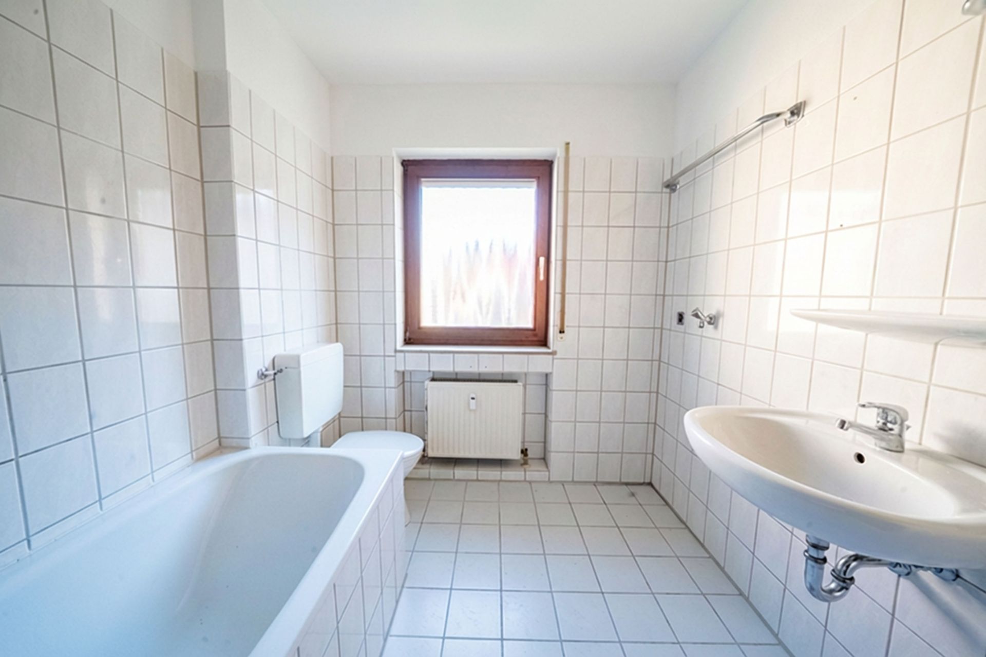 Helles Badezimmer mit Badewanne, WC, Waschbecken und Fenster, weiße Fliesen an Wänden und Boden.