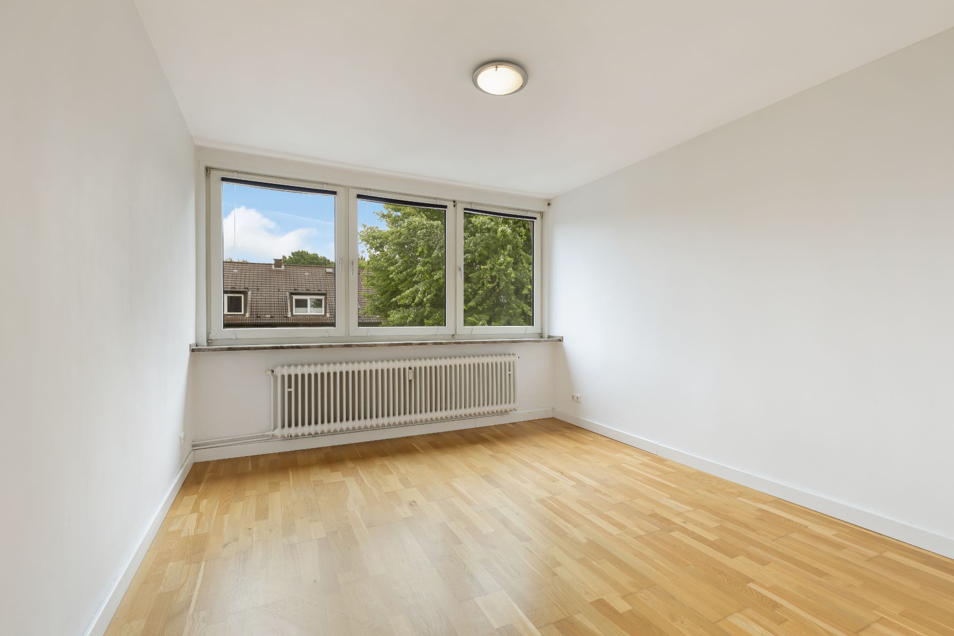 Leerer Raum mit Holzboden, weißen Wänden und großem Fenster mit Blick auf Bäume.