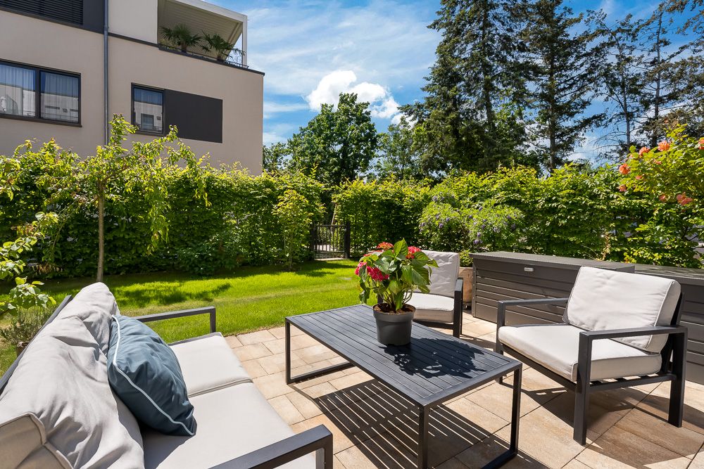 Moderne Terrasse mit Sitzgruppe, Tisch und Pflanze, umgeben von grünem Garten und Hecke.