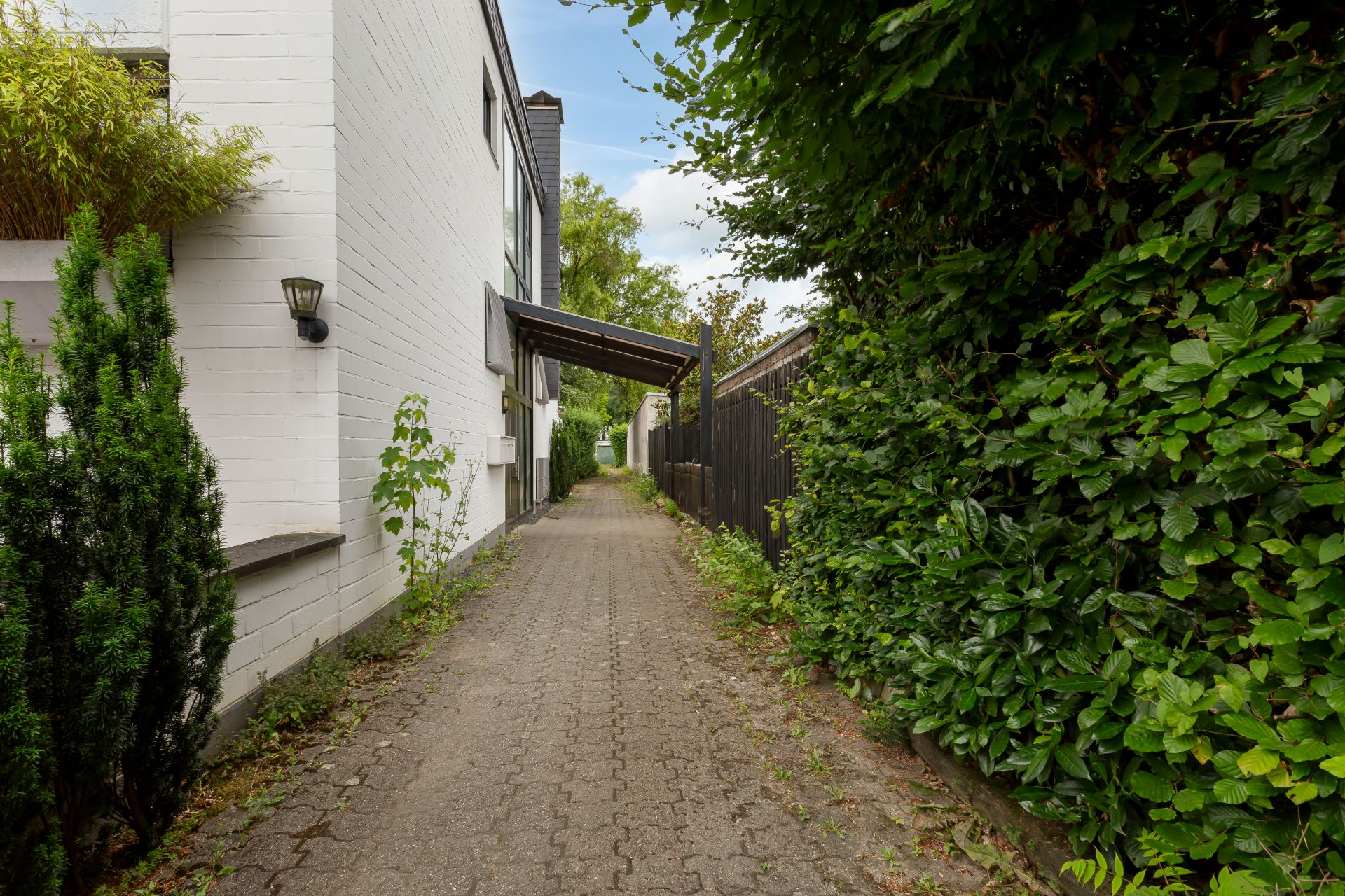 Gepflasterter Weg neben einem weißen Wohnhaus mit Büschen und Hecke.