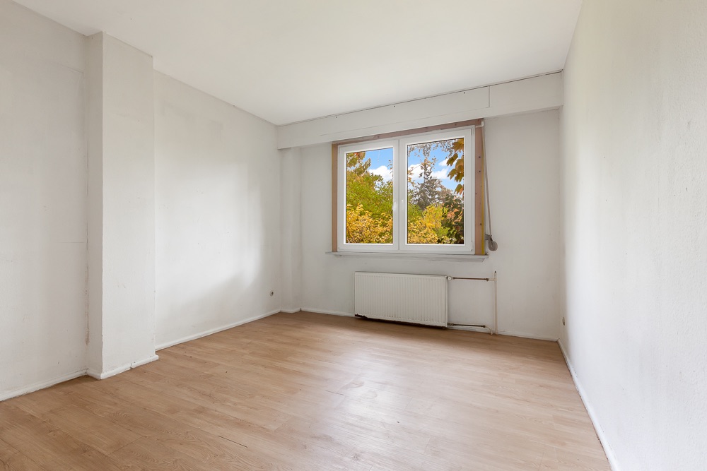 Leeres Zimmer mit Holzfußboden, weißer Wand und Fenster mit Blick auf Bäume.