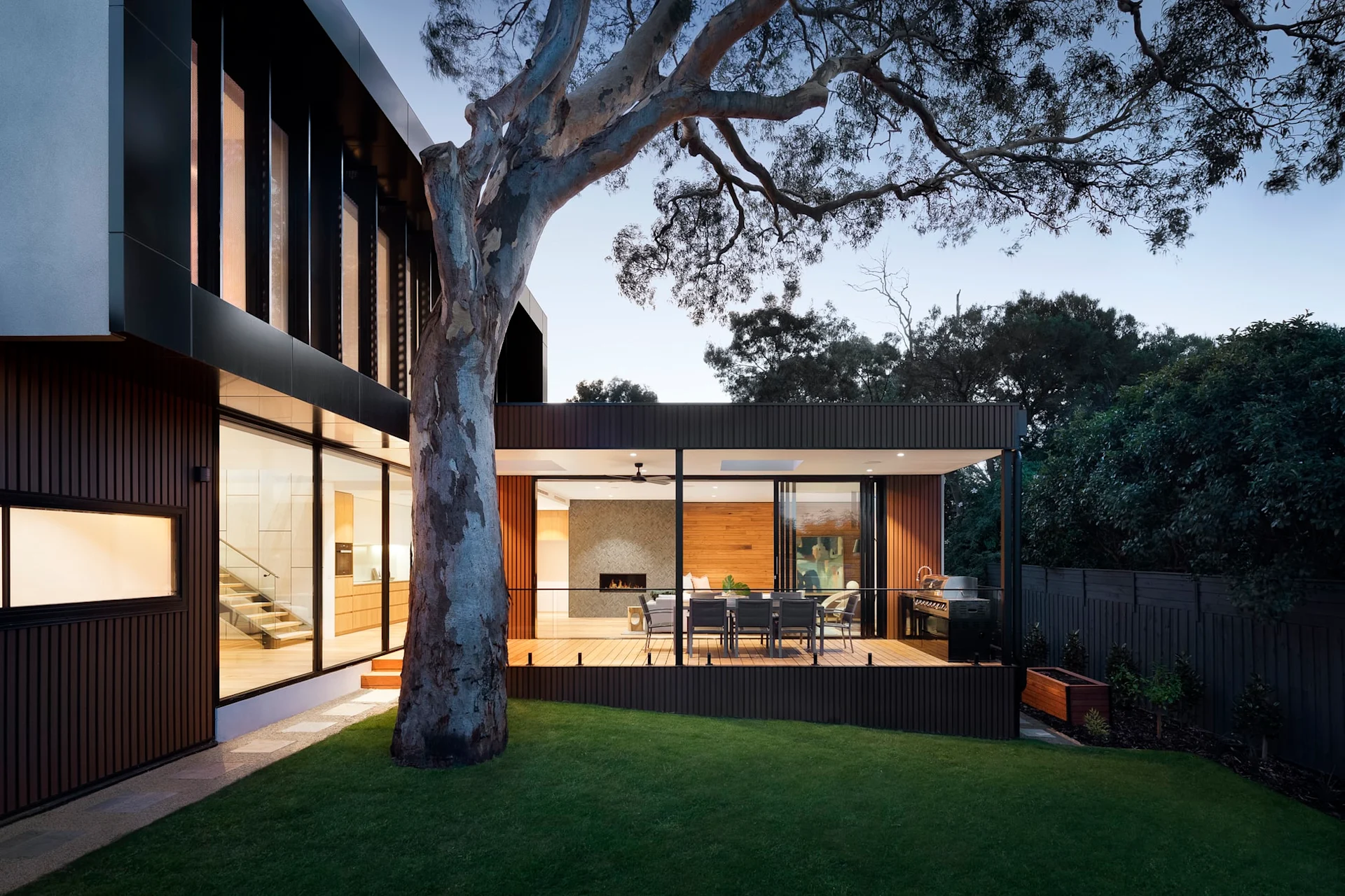 Modernes Wohnhaus mit großer Terrasse, Essbereich und Grill im Garten, Baum im Vordergrund.