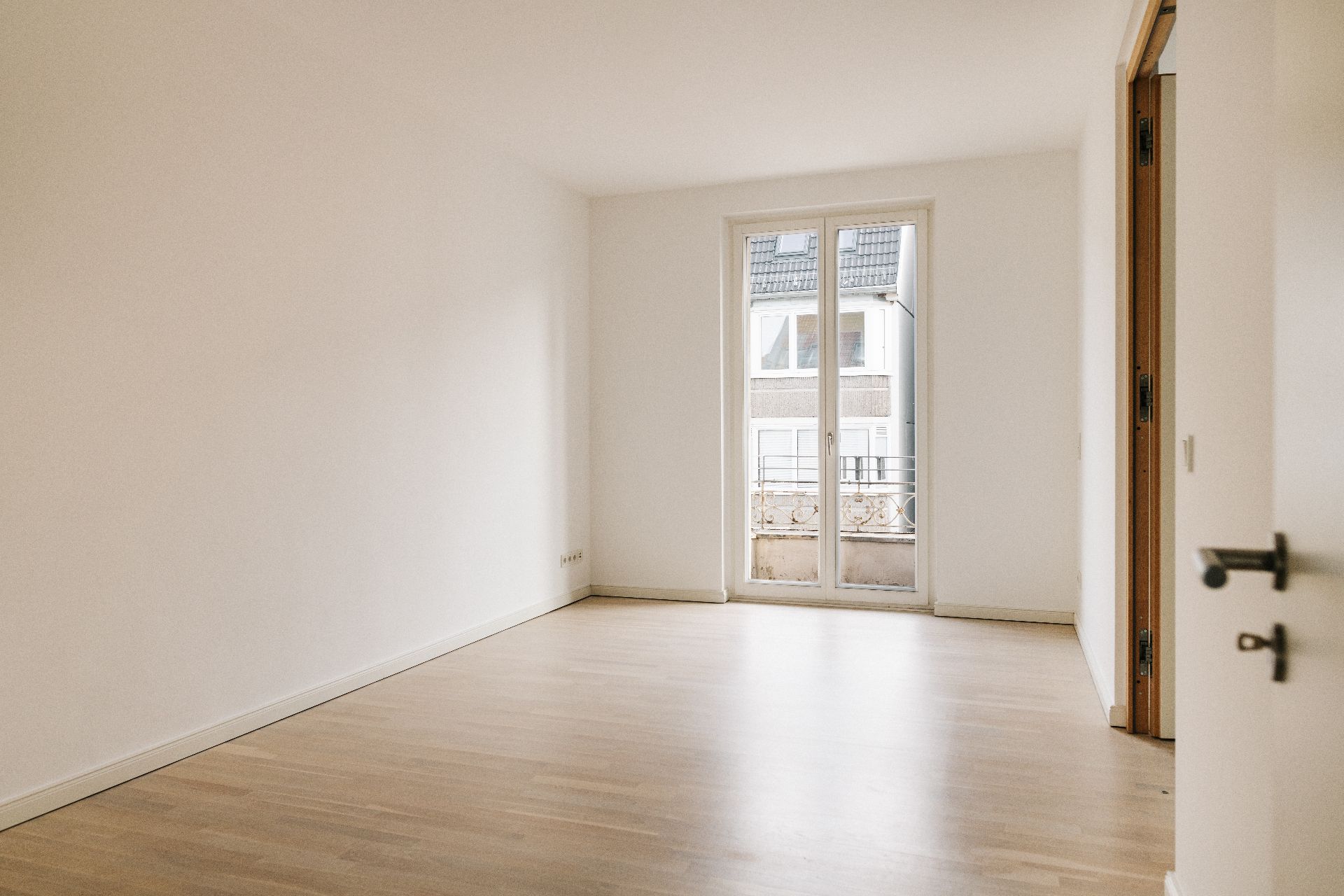 Helles, leeres Zimmer mit Parkettboden und bodentiefem Fenster zu einem kleinen Balkon.