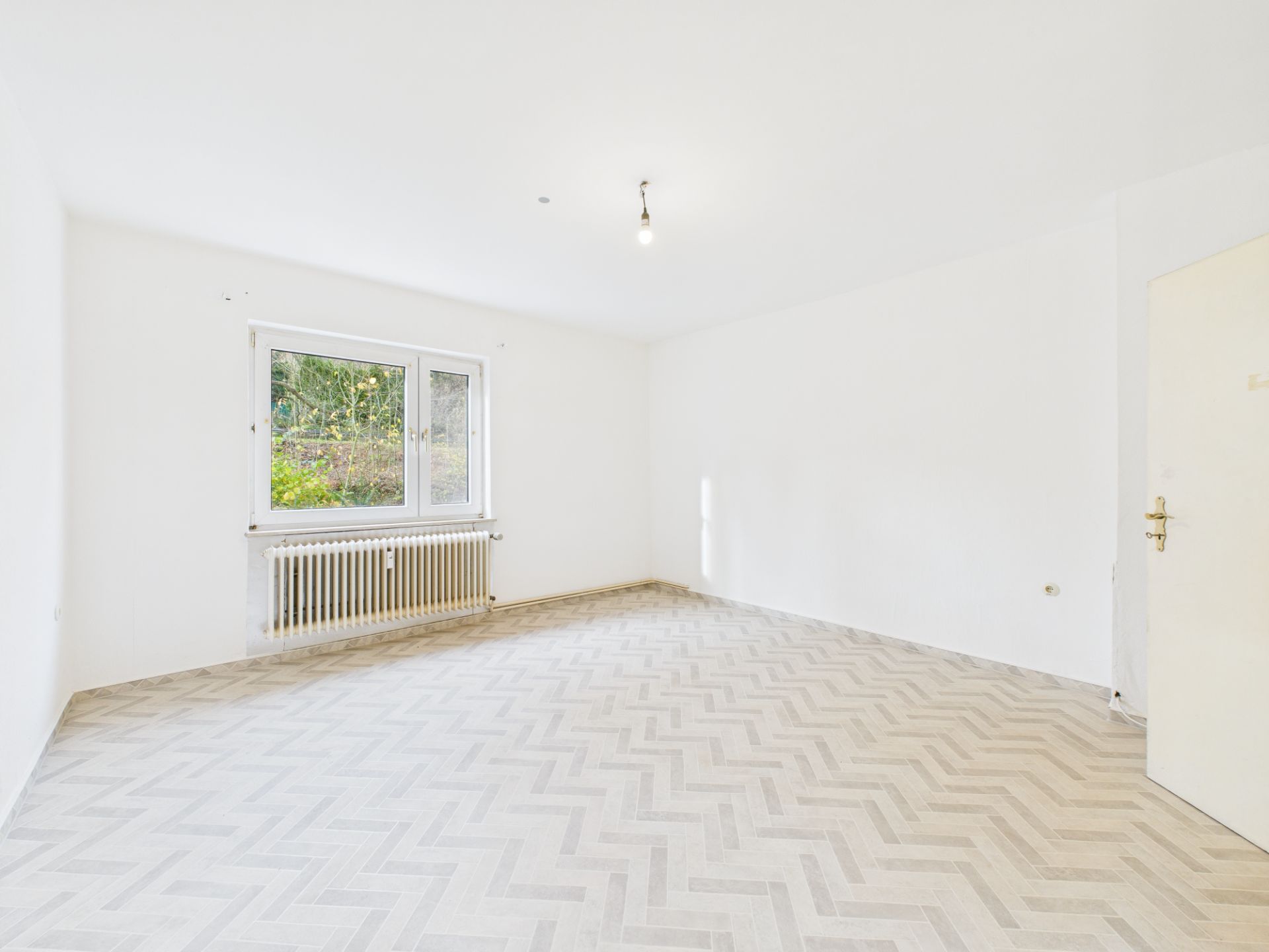 Leerer, heller Raum mit weißer Wand, großem Fenster und Radiator, Zickzack-Bodenmuster.