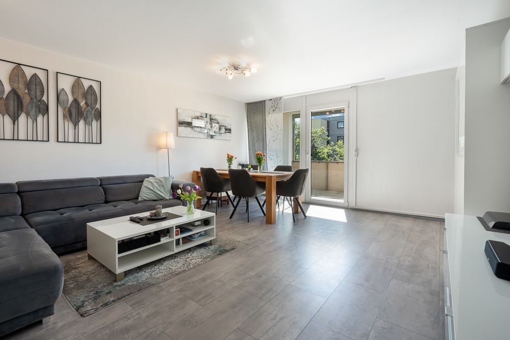 Helles Wohnzimmer mit Ecksofa, Couchtisch, Esstisch und Zugang zum Balkon.