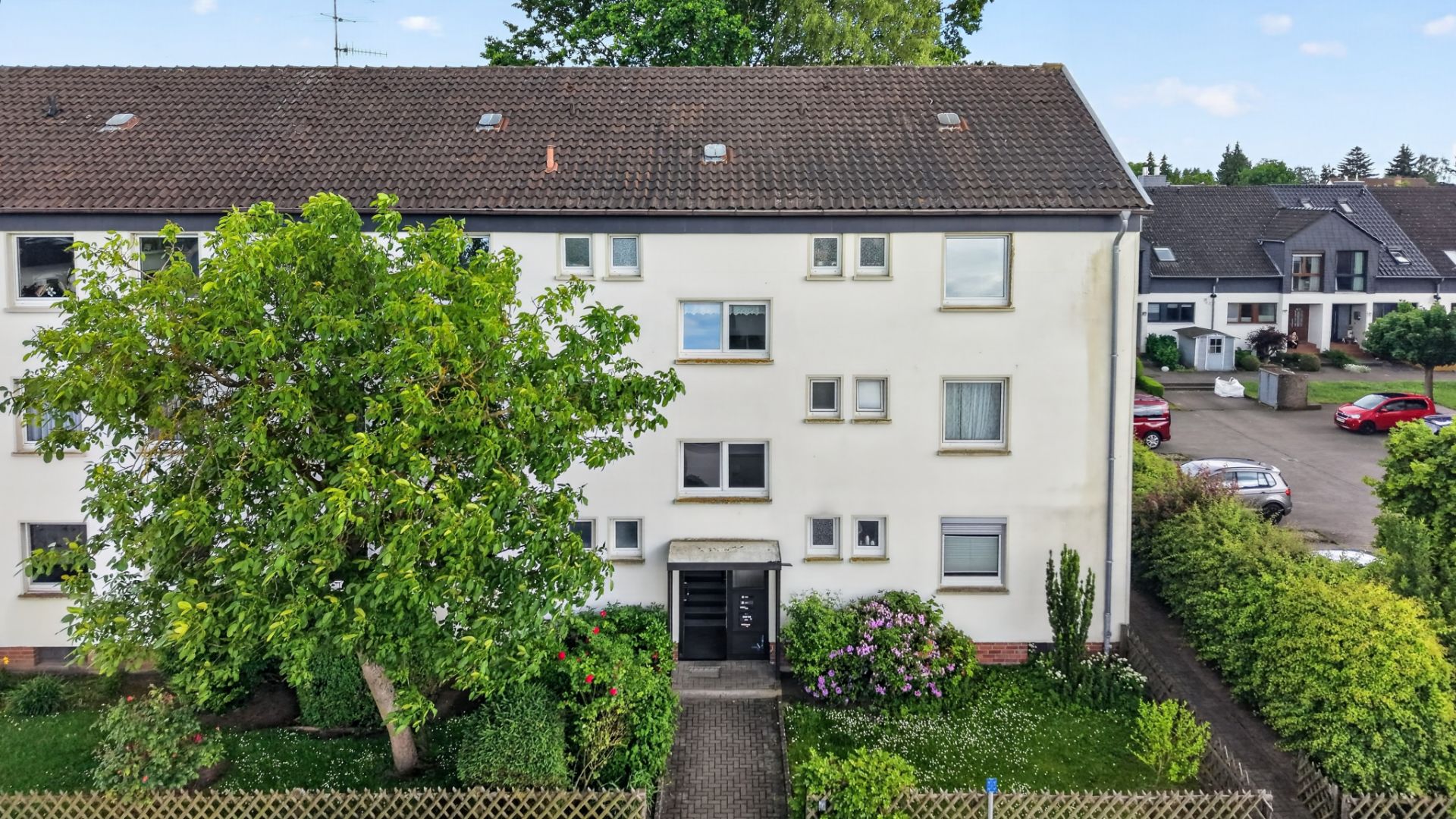 Dreistöckiges Wohnhaus mit Vorgarten, Bäumen und Hecke, Eingang in der Mitte.