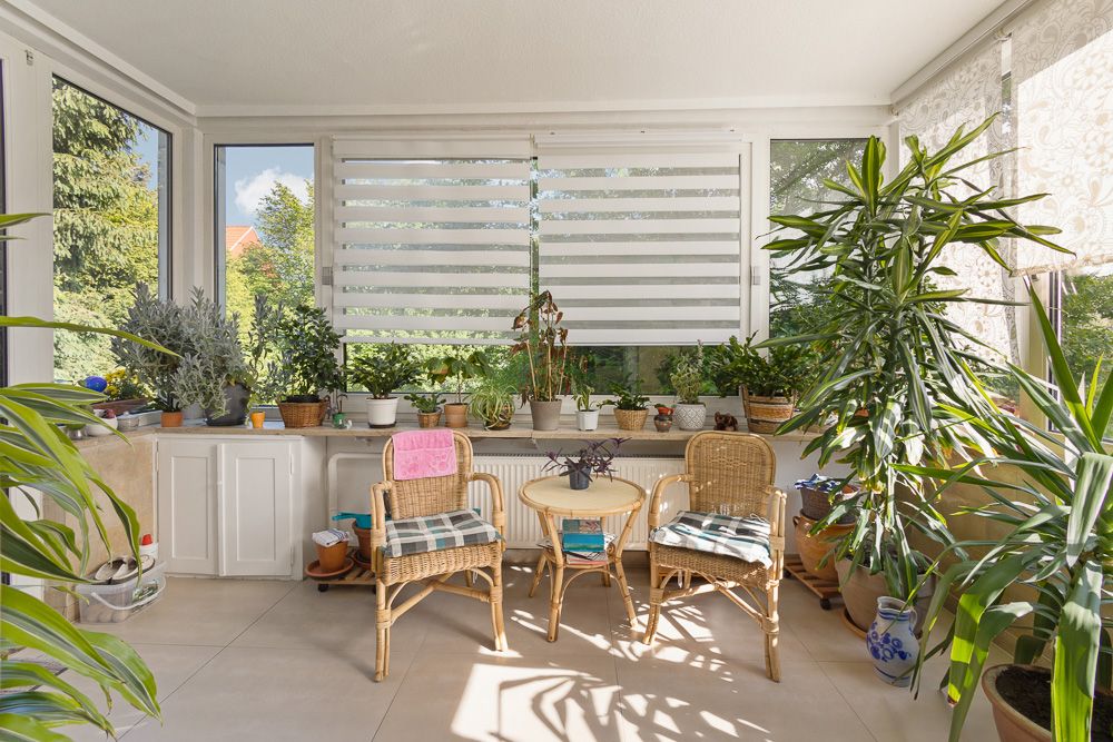 Heller Wintergarten mit zwei Korbstühlen, Tisch und vielen Topfpflanzen am Fenster.