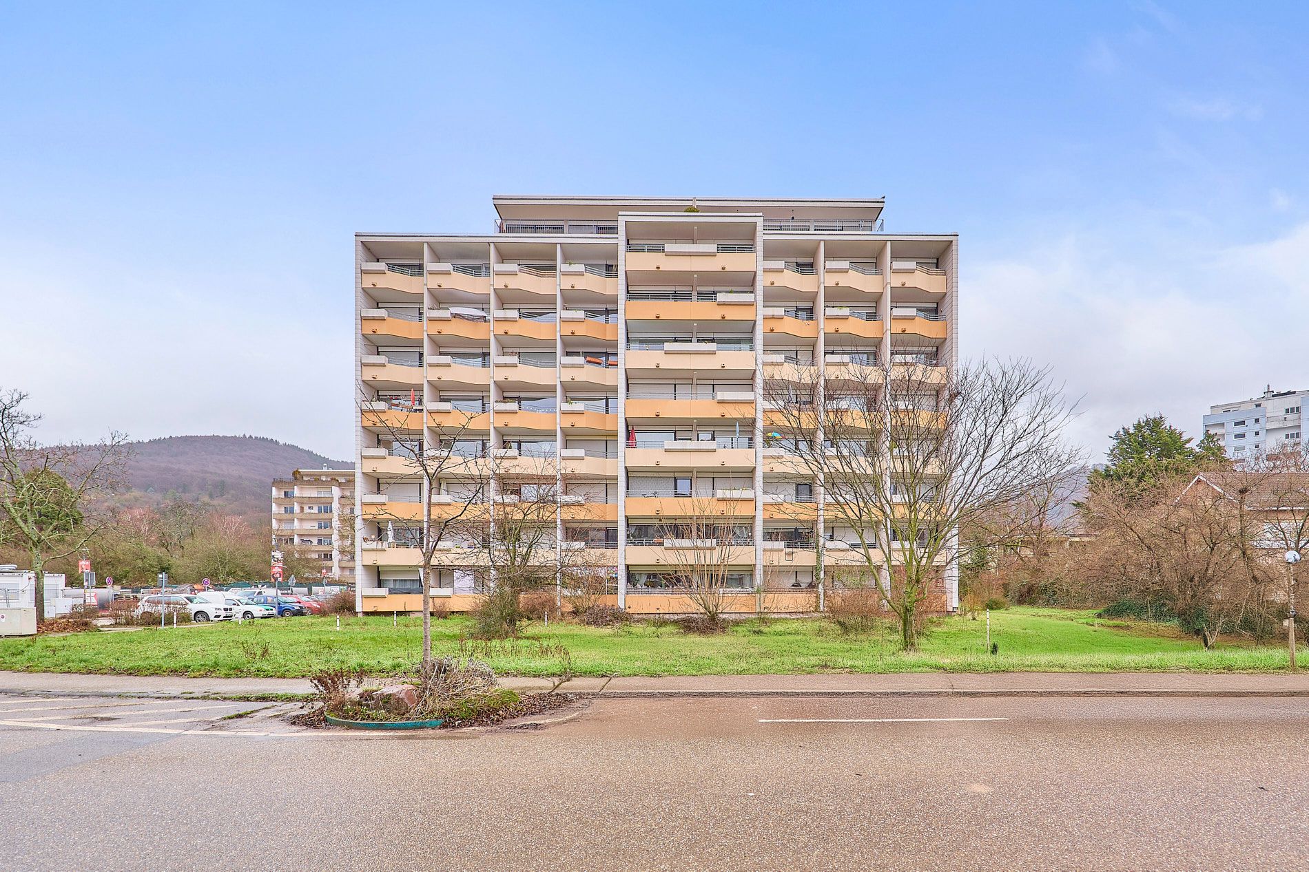Mehrstöckiges Wohngebäude mit vielen Balkonen, davor eine Straße und etwas Grünfläche.