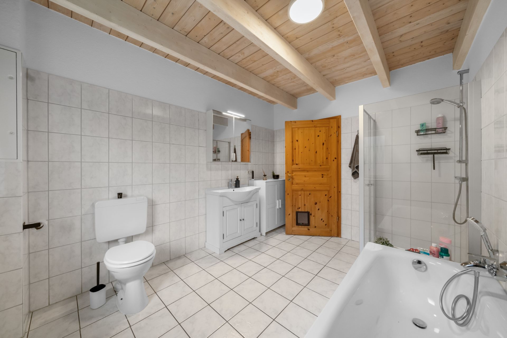 Modernes Badezimmer mit Badewanne, Dusche, WC und Holztür, gefliestem Boden und Holzbalkendecke.