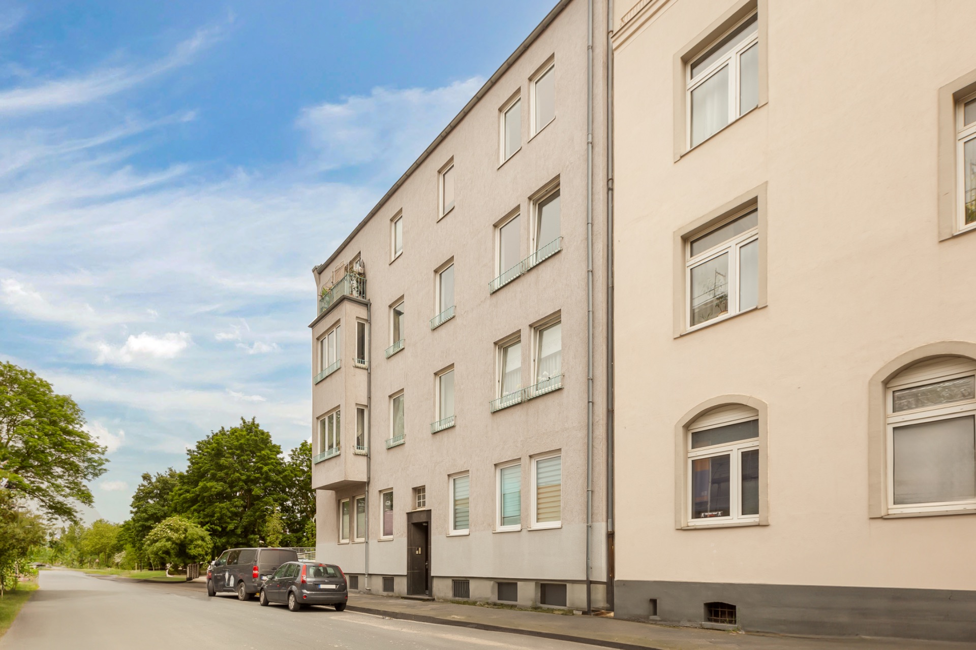 Mehrfamilienhaus mit mehreren Fenstern und parkenden Autos am Straßenrand.