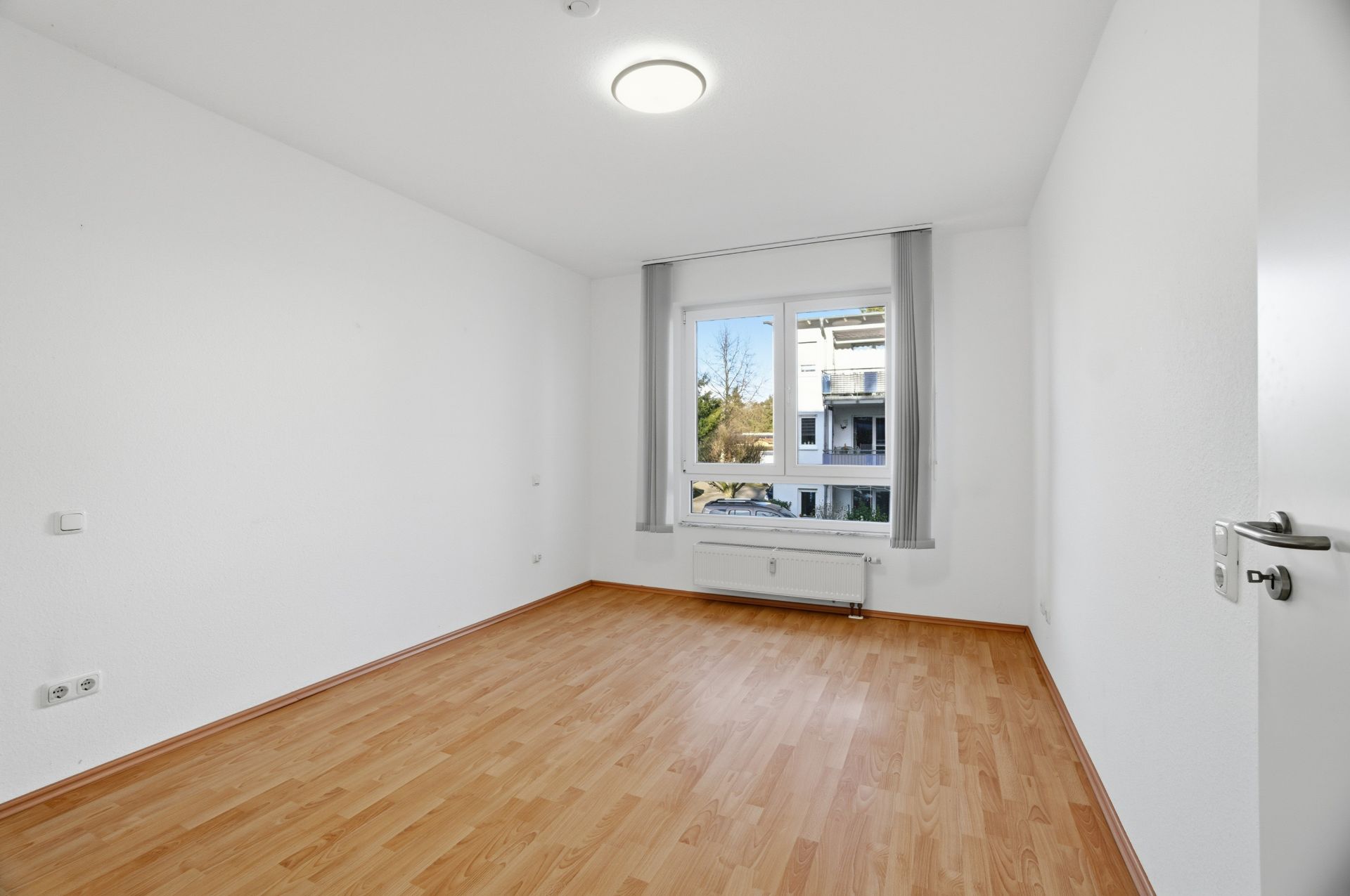 Leeres Zimmer mit Holzfußboden, weißem Wänden und großem Fenster mit Blick nach draußen.