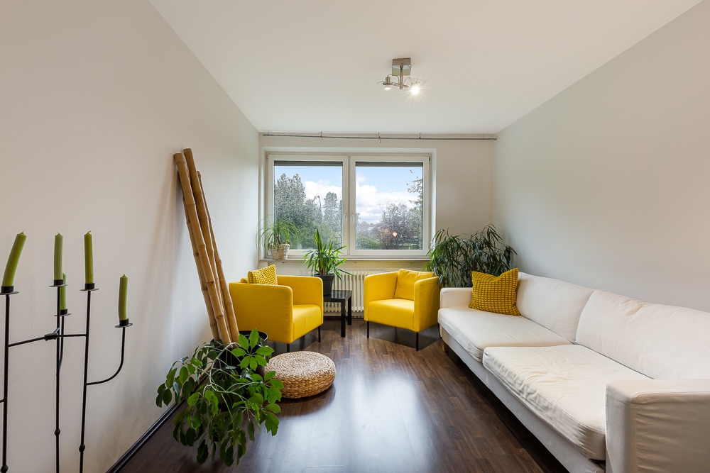 Helles Wohnzimmer mit weißem Sofa, zwei gelben Sesseln, Pflanzen und großem Fenster.