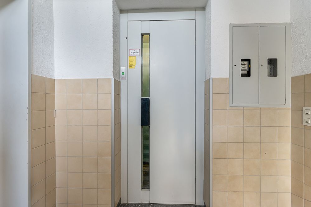 Weißer Aufzug mit geschlossener Tür zwischen beige gefliesten Wänden im Wohnhausflur.