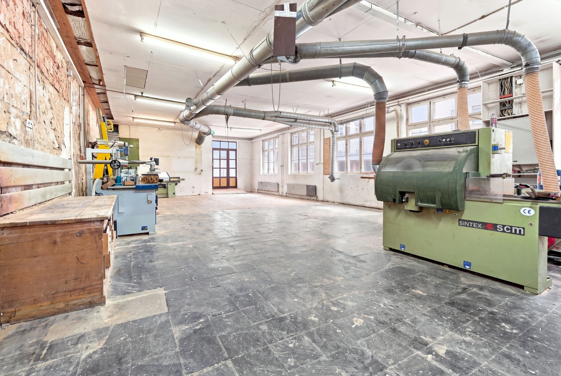 Großer Werkstattraum mit Maschinen, Werkbänken und sichtbaren Lüftungsrohren an der Decke.