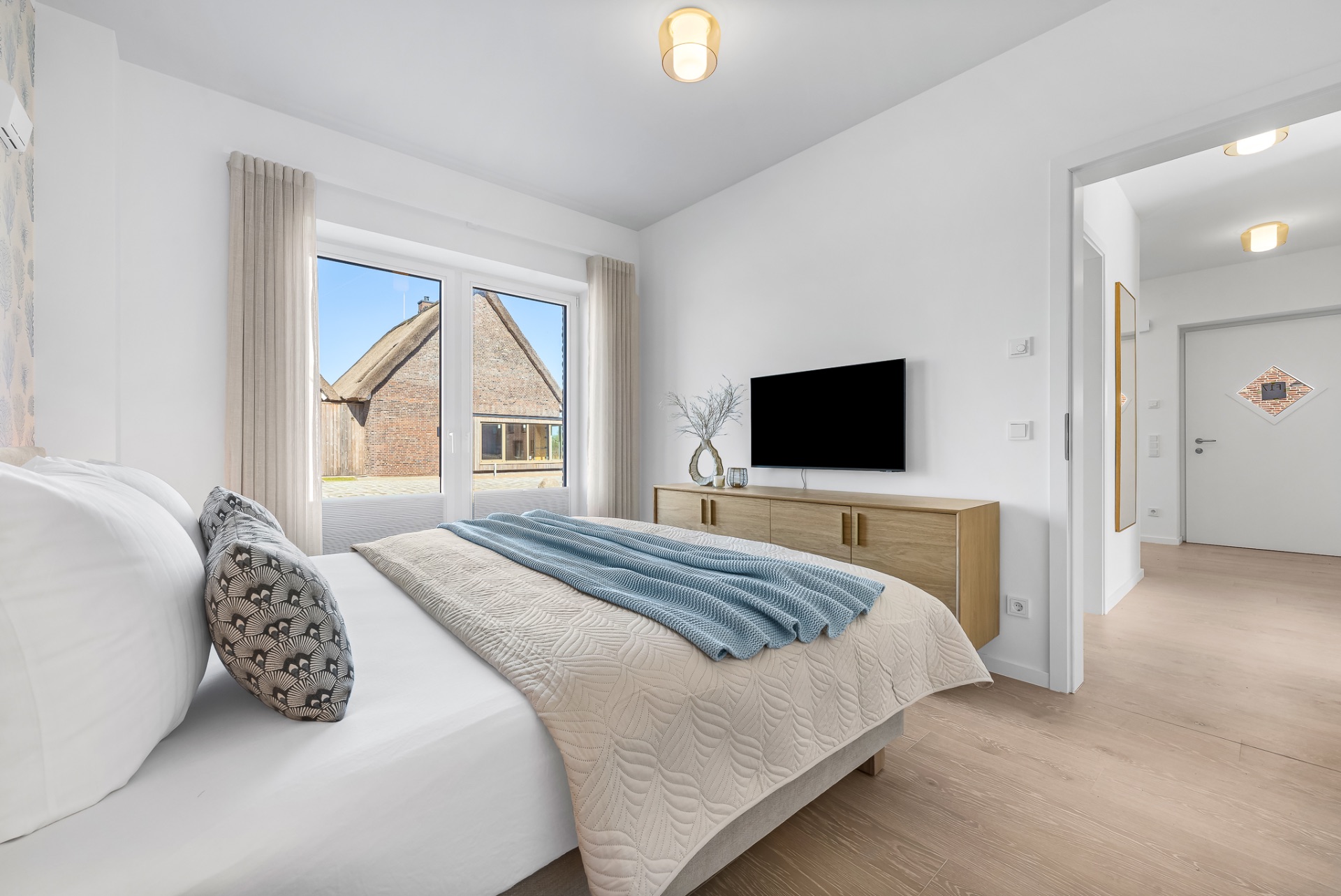 Helles Schlafzimmer mit Doppelbett, TV, Kommode und großem Fenster mit Blick auf ein Haus.