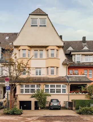 Mehrstöckiges Wohnhaus mit Erker, Auffahrt und geparktem Auto, daneben weitere Wohnhäuser.