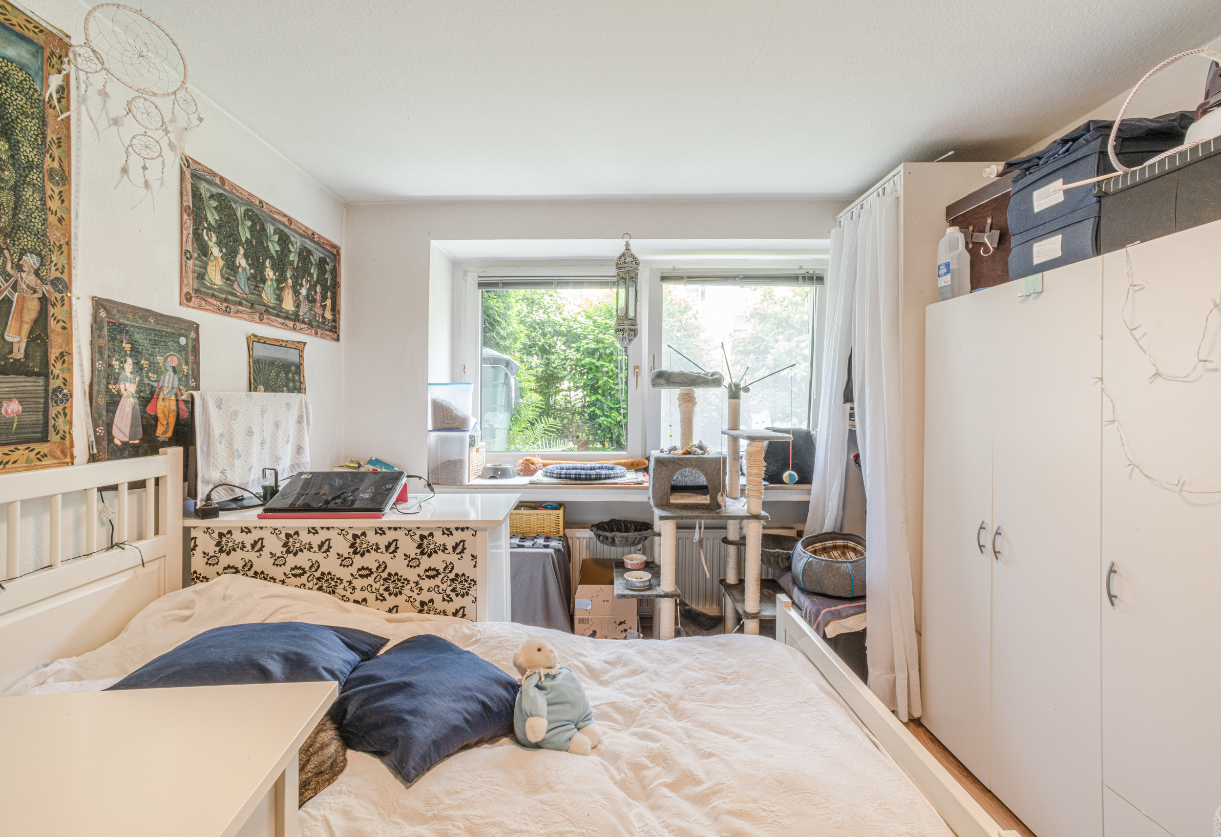Helles Schlafzimmer mit Bett, Schrank, Schreibtisch, Katzenbaum und Dekoration an den Wänden.