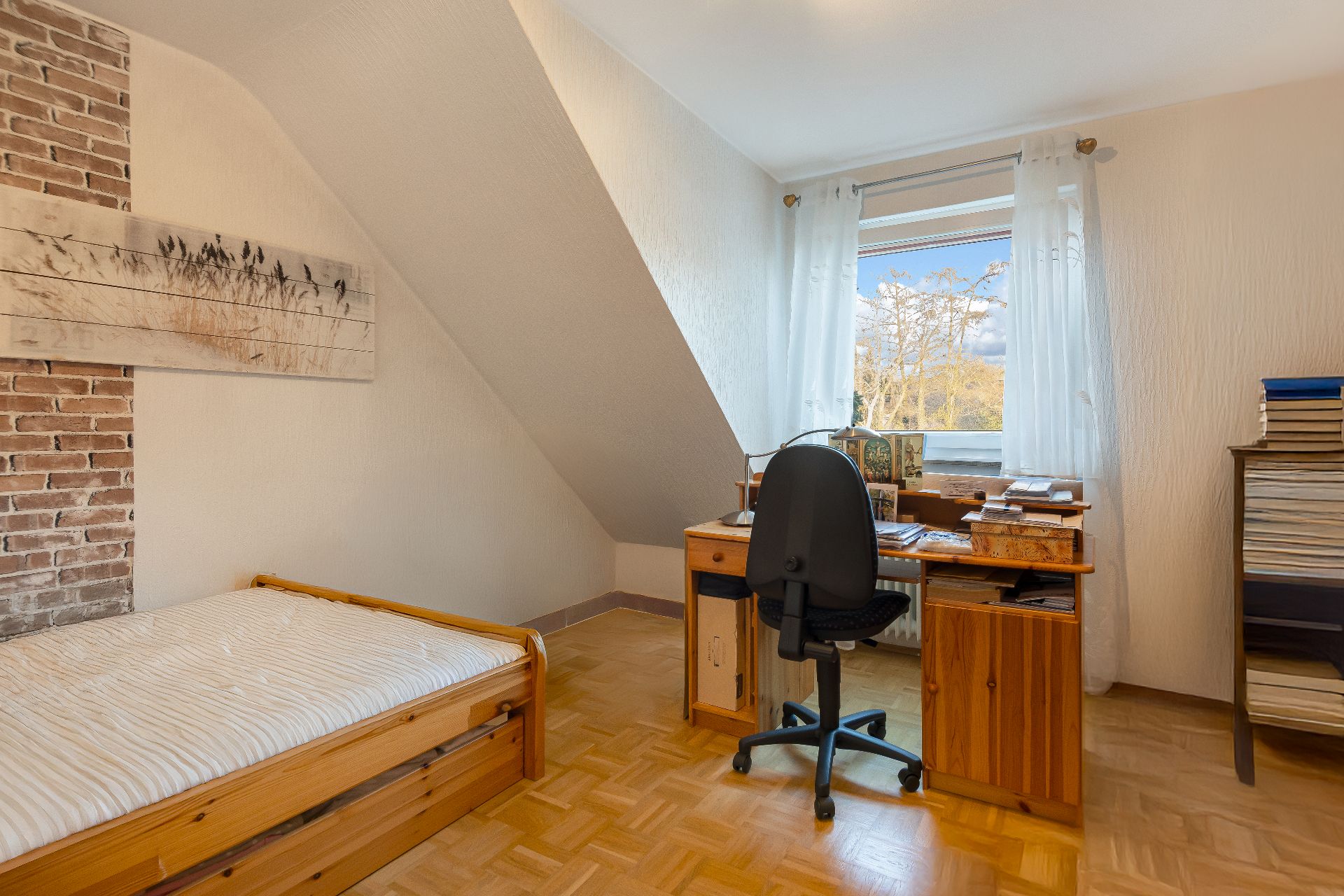 Kleines Zimmer mit Holzbett, Schreibtisch, Stuhl und Fenster mit Blick nach draußen.